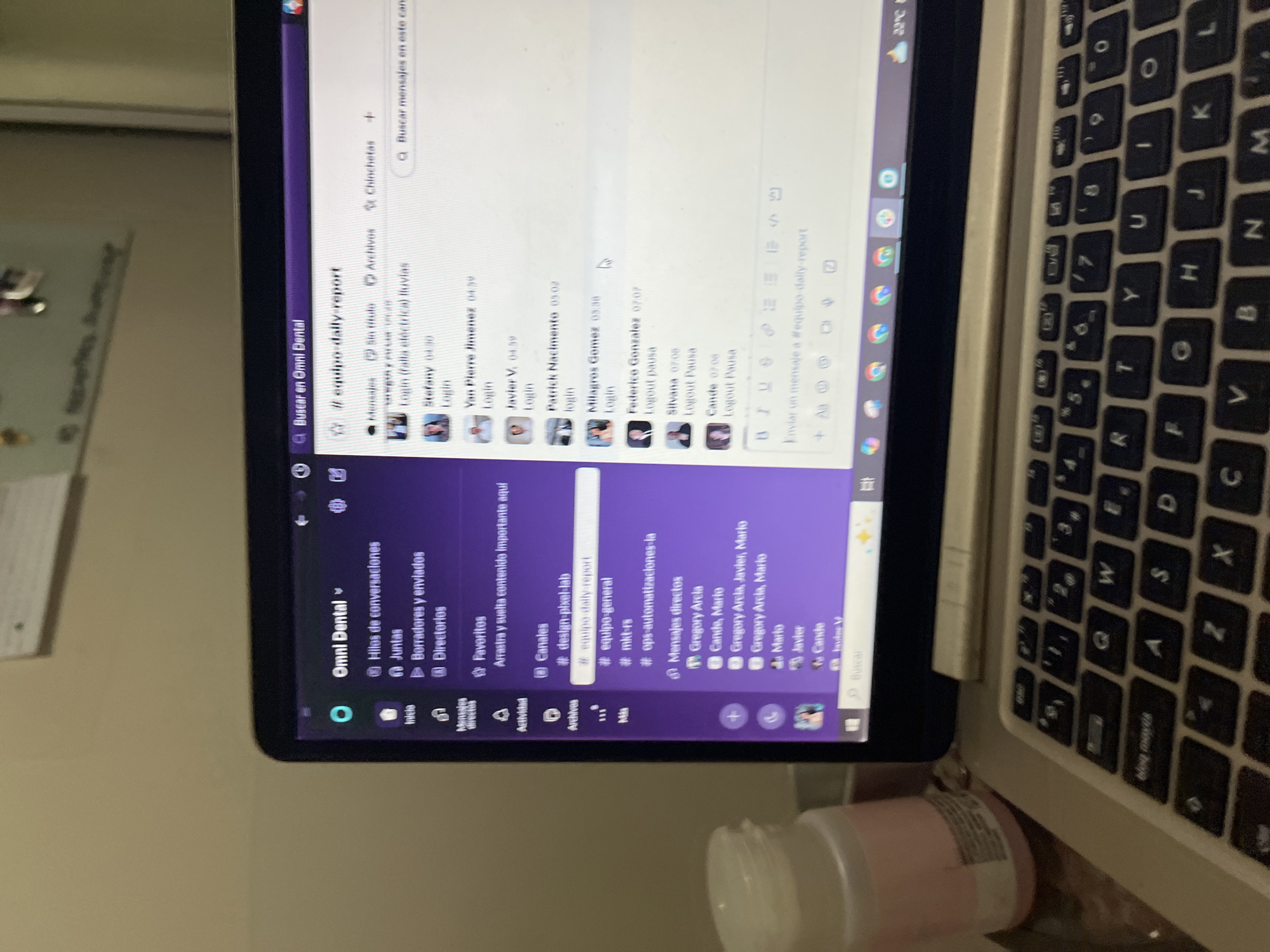 An eye-level, tilted shot captures a laptop displaying a communication platform, likely in an indoor setting in San Luis, Peru. The laptop screen is the central focus, showcasing what appears to be a Slack-like application with a purple sidebar and a white main chat window.
On the screen, the application is branded "Omni Dental." The left sidebar organizes content under categories like "Hilos de conversaciones" (Conversation threads), "Juntas" (Meetings), "Borradores y enviados" (Drafts and sent), and "Directorios." Several channels are listed, including "#design-pixel-lab," "#equipo-daily-report" (team daily report), "#equipo-general," and "#ops-automatizaciones-la." Direct messages to various individuals, such as Gregory Arcia, Cande, Mario, and Javier, are also visible.
The main chat window is focused on the "#equipo-daily-report" channel, showing a series of messages indicating team members' activity. Entries like "Stefany 04:30 Login," "Van Pierre Jimenez 04:39 Login," "Patrick Nacimento 03:02 Login," and "Milagros Gomez 03:38 Login" are visible, along with "Logout pausa" entries for Federico Gonzalez (07:07), Silvana (07:08), and Cande (07:08). A search bar at the top right reads "Buscar mensajes en este canal," and a small weather icon indicates 22°C.
The right side of the image reveals part of the laptop's physical keyboard, which features a Spanish QWERTY layout with keys like 'Ñ' and "bloq mayús" (caps lock). In the immediate foreground, a light pink container or bottle is partially visible. The background shows a plain, light-colored wall. The overall scene suggests a work environment where team members communicate and track daily activities.