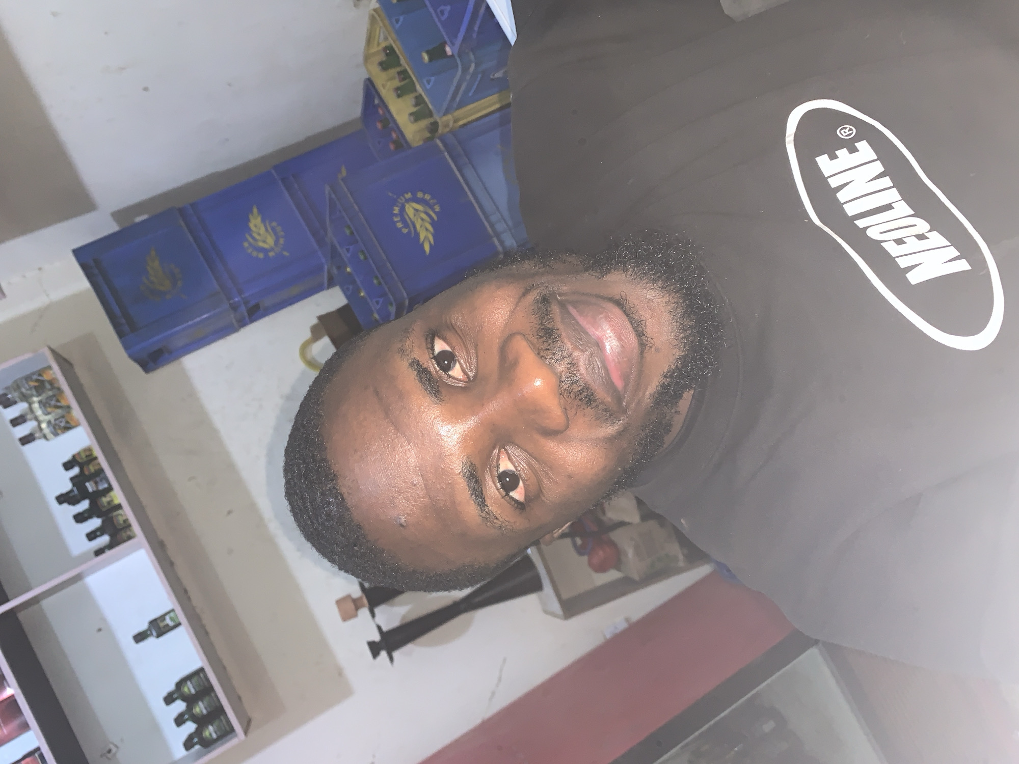 The image shows a close-up selfie of a Black man with short, dark, curly hair and a beard, smiling directly at the camera. He is wearing a black t-shirt with a white oval logo that reads "NEOLINE". The background is somewhat cluttered. To the left, there are shelves with various small bottles, possibly liquor. To the right, there are stacks of blue plastic crates, some containing bottles. The setting appears to be an indoor space, possibly a shop or storage area, in New Stadium, Nigeria. The lighting is dim, creating some shadows.