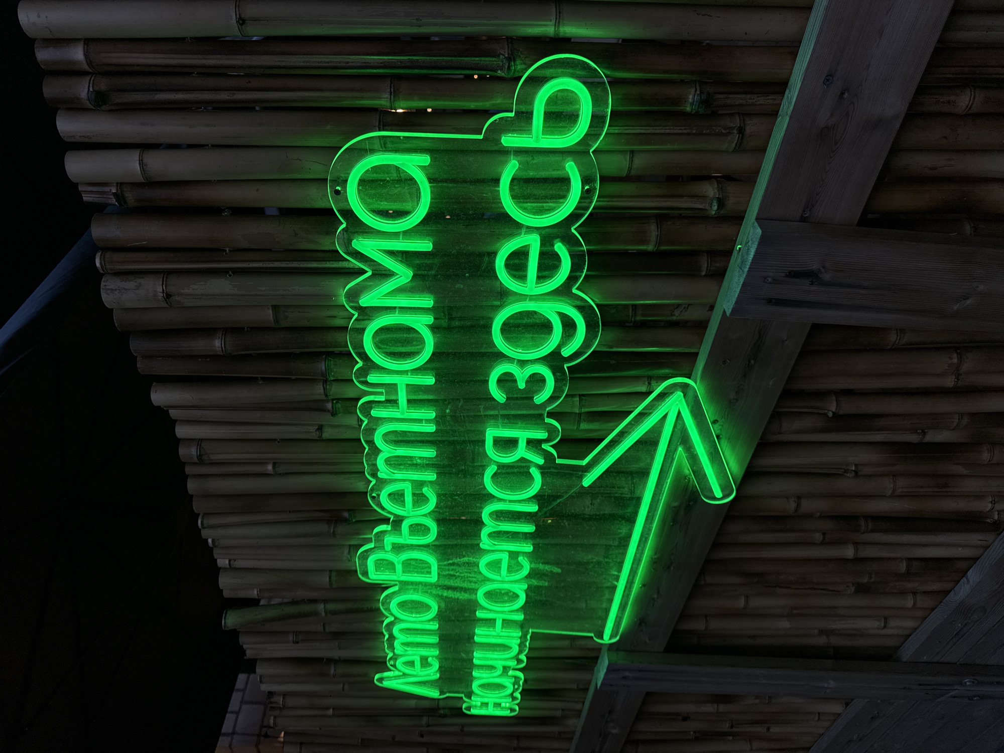 A neon sign with the Russian text "Лето Вьетнама начинается здесь" (Summer Vietnam starts here) glows in green against a backdrop of bamboo and wood. The sign includes an arrow pointing upwards. The scene is likely located in Moscow, Russia, based on the Cyrillic script used.