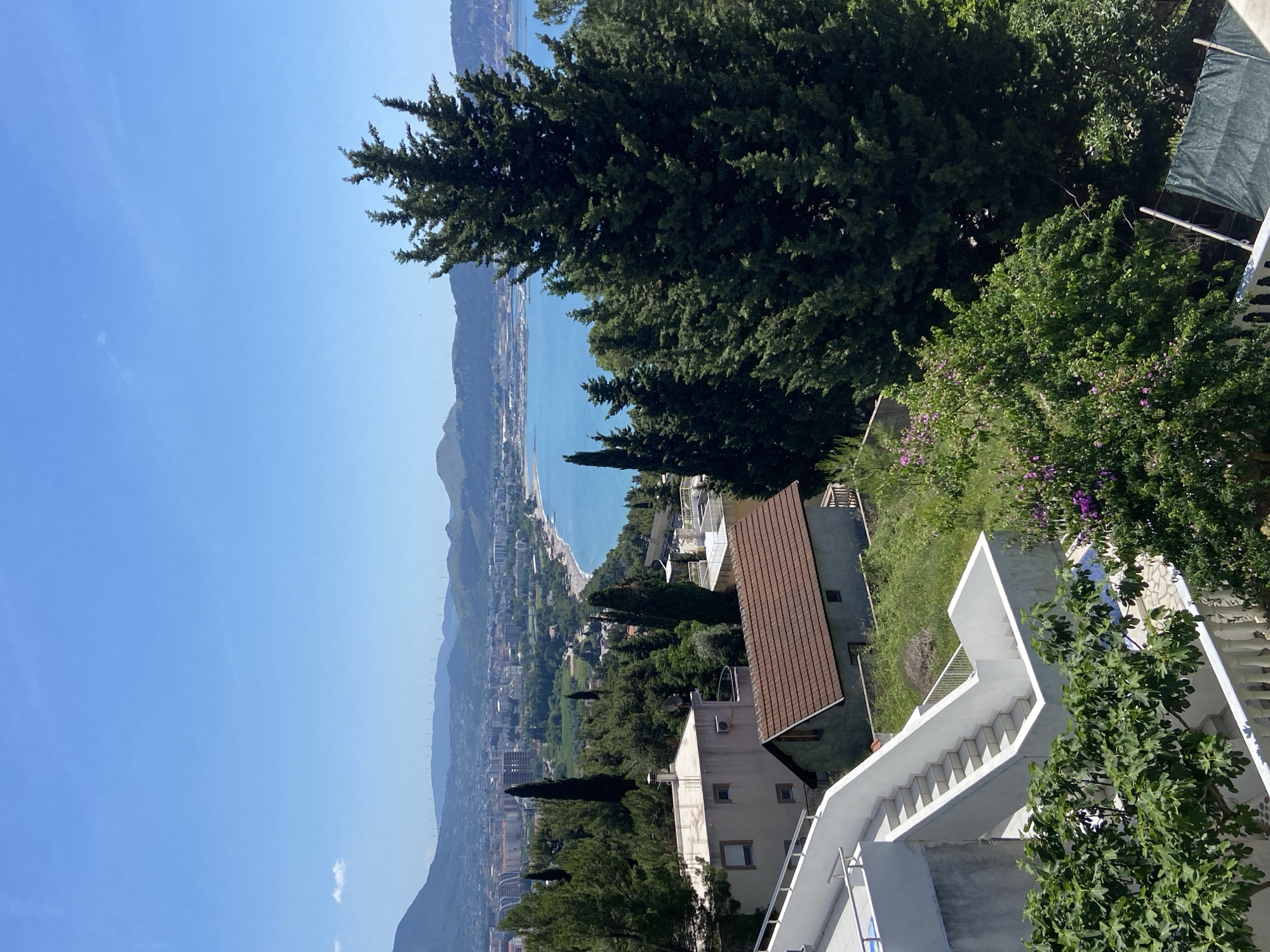This is an eye-level, outdoor photograph of Šušanj, Montenegro, on a sunny day. In the foreground are white stairs with a grey railing leading up the side of a building, with lush trees framing the right side. A two-story house with a brown roof and white siding is visible, with a taller tree behind it. In the background, a beach town stretches along the coastline, with a mountain range behind it. The sky is a clear blue.