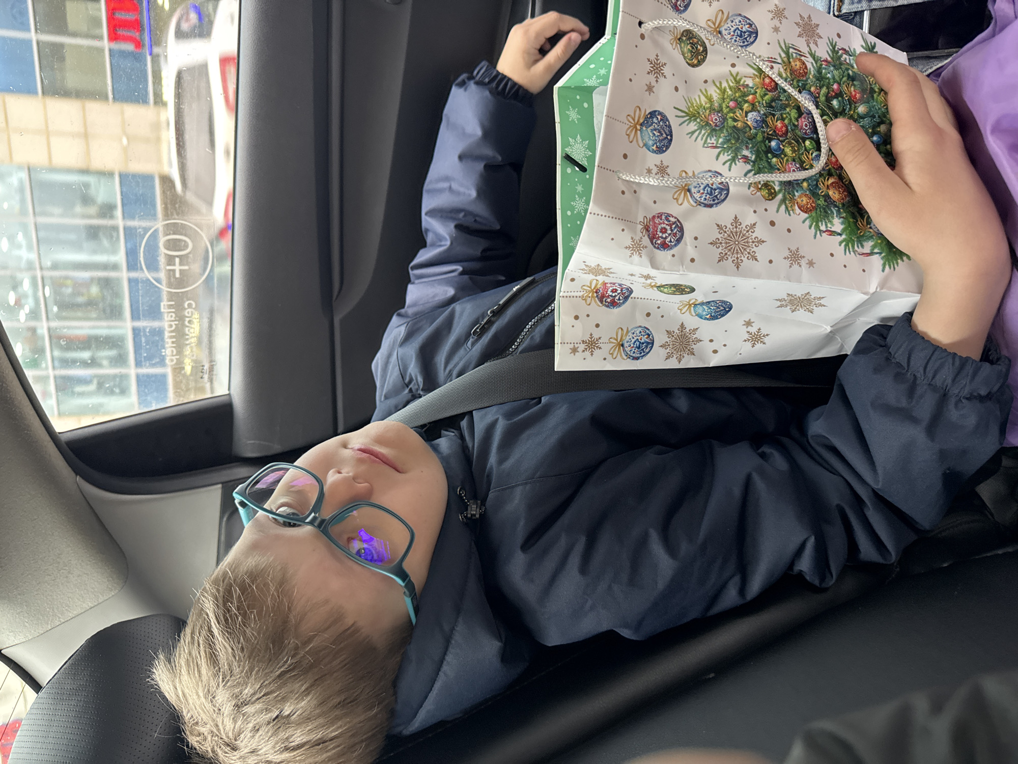 A young boy wearing glasses and a navy blue jacket is lying in the back seat of a car, looking at a gift bag. The gift bag is white with a Christmas tree design and snowflake decorations. The boy is wearing a seatbelt. The car window is visible, showing a building and other cars outside, possibly in the city of Perm, Russia. The window displays the "+" sign for "Children."