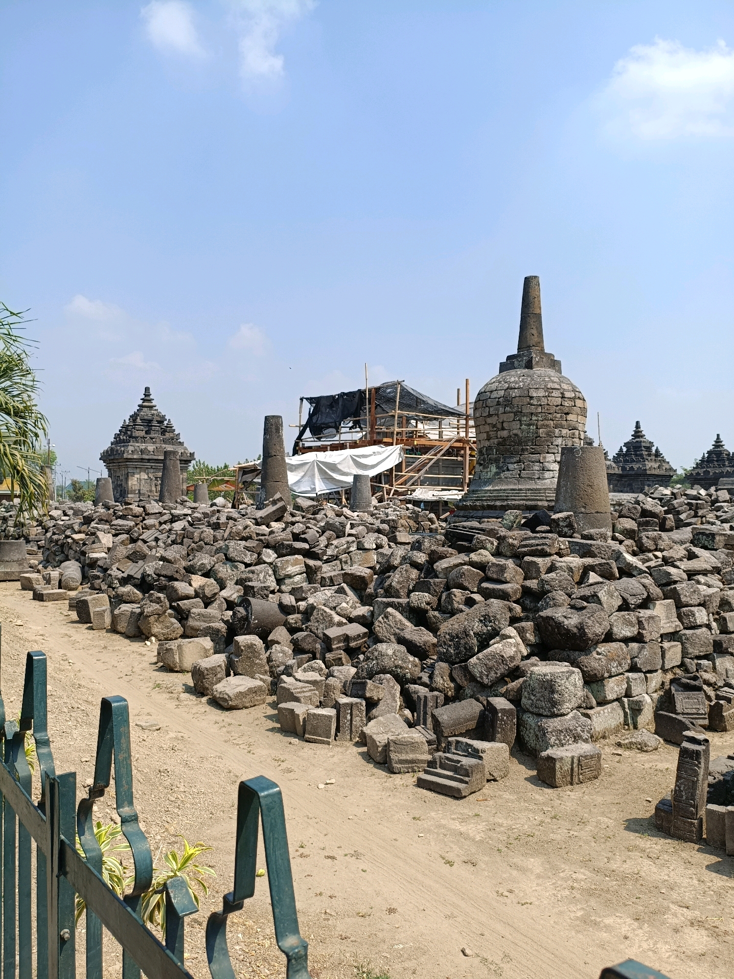 The image depicts the Plaosan Temple complex, a historical Buddhist temple located in Bugisan, Indonesia. The foreground features a green metal fence, beyond which lies a sandy path. The central and background areas are dominated by numerous stone ruins, piled high and scattered across the ground, indicating past construction and potential reconstruction efforts. Several stone stupas, characteristic of Buddhist architecture, are visible, with one prominent stupa in the mid-ground surrounded by debris. In the background, more temple structures with tiered roofs are discernible, suggesting the scale of the complex. Construction scaffolding and tarps are present, hinting at ongoing restoration or preservation work. The sky is a clear blue with a few white clouds, and the overall lighting suggests a bright, sunny day. No people are visible in the image.