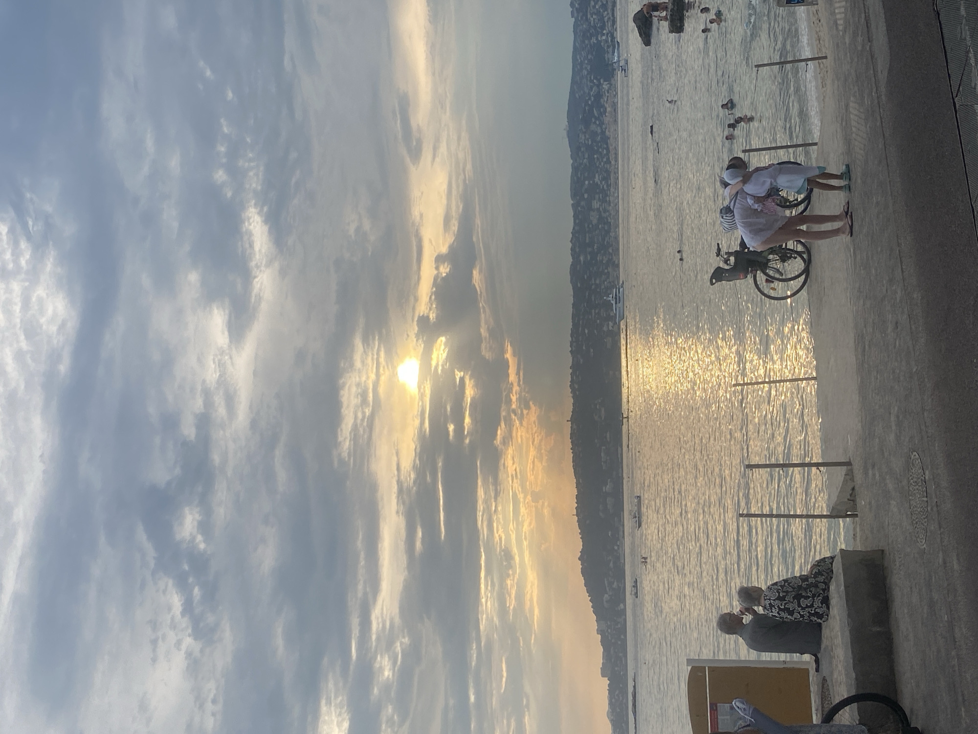 In Antibes, France, at sunset, a tranquil scene unfolds by the Mediterranean Sea. The sky is a canvas of dramatic clouds, with the sun's rays piercing through, casting a golden shimmer on the water. In the foreground, two elderly individuals are seated on a concrete bench, facing the sea, with one appearing to drink from a cup. To their right, a woman dressed in white bends over a child, also in white, near a bicycle. In the background, boats dot the water, and the coastline is dotted with buildings. Several people are seen swimming and wading in the sea, enjoying the last moments of daylight. The overall atmosphere is one of peaceful relaxation and natural beauty.