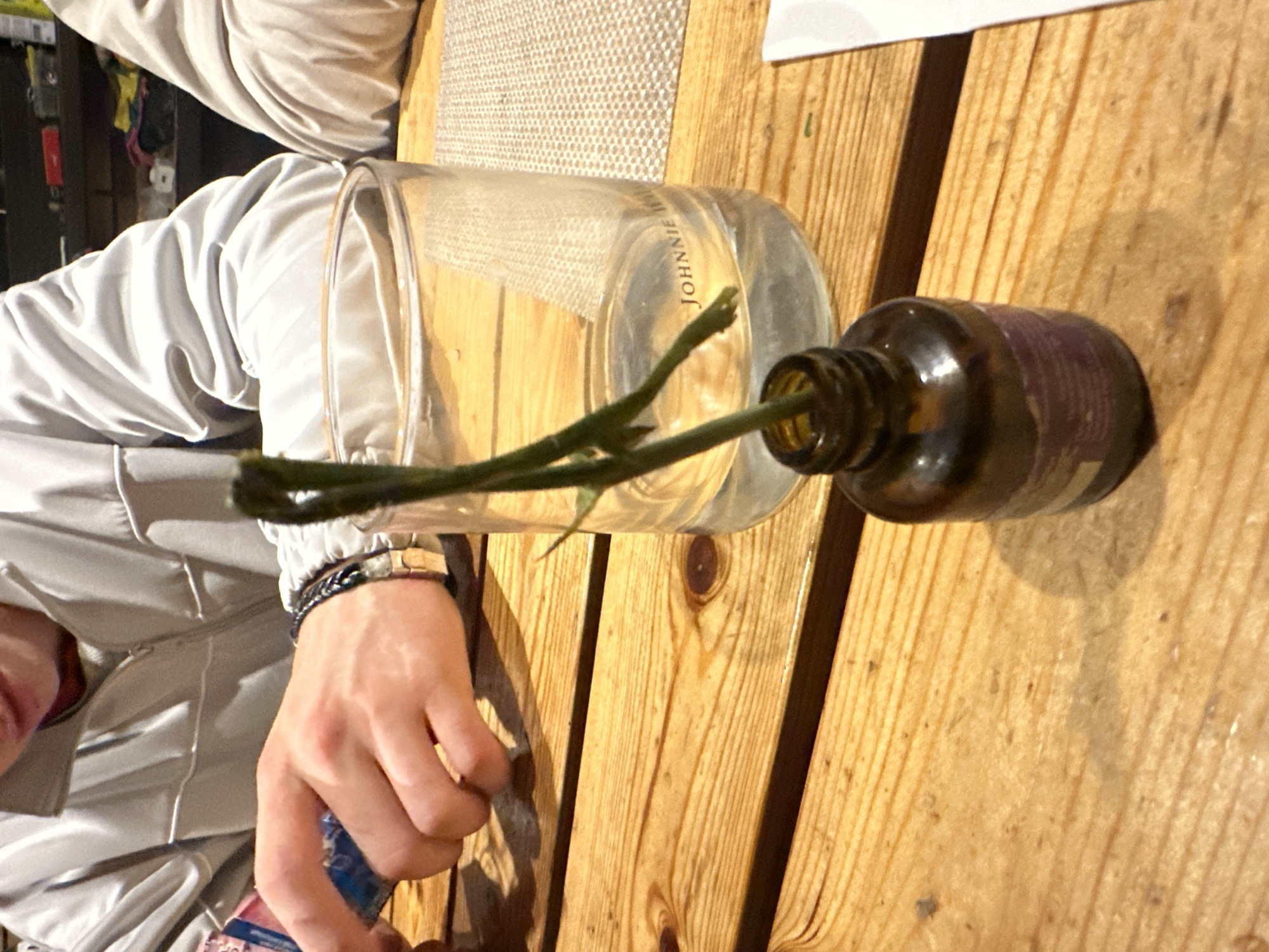The image depicts a close-up view of a wooden table, likely in a restaurant or home setting, as suggested by the presence of a glass with "JOHNNIE" etched on it, a small dark brown bottle with a purple label, and a green plant stem emerging from the bottle. Two individuals are partially visible in the background, one with their hands on the table interacting with what appears to be a package, and another person sitting to the right. The lighting suggests an indoor environment, possibly with ambient or dim lighting. The exact location in Востец Снт, Russia, cannot be determined from the visual information provided in the image.