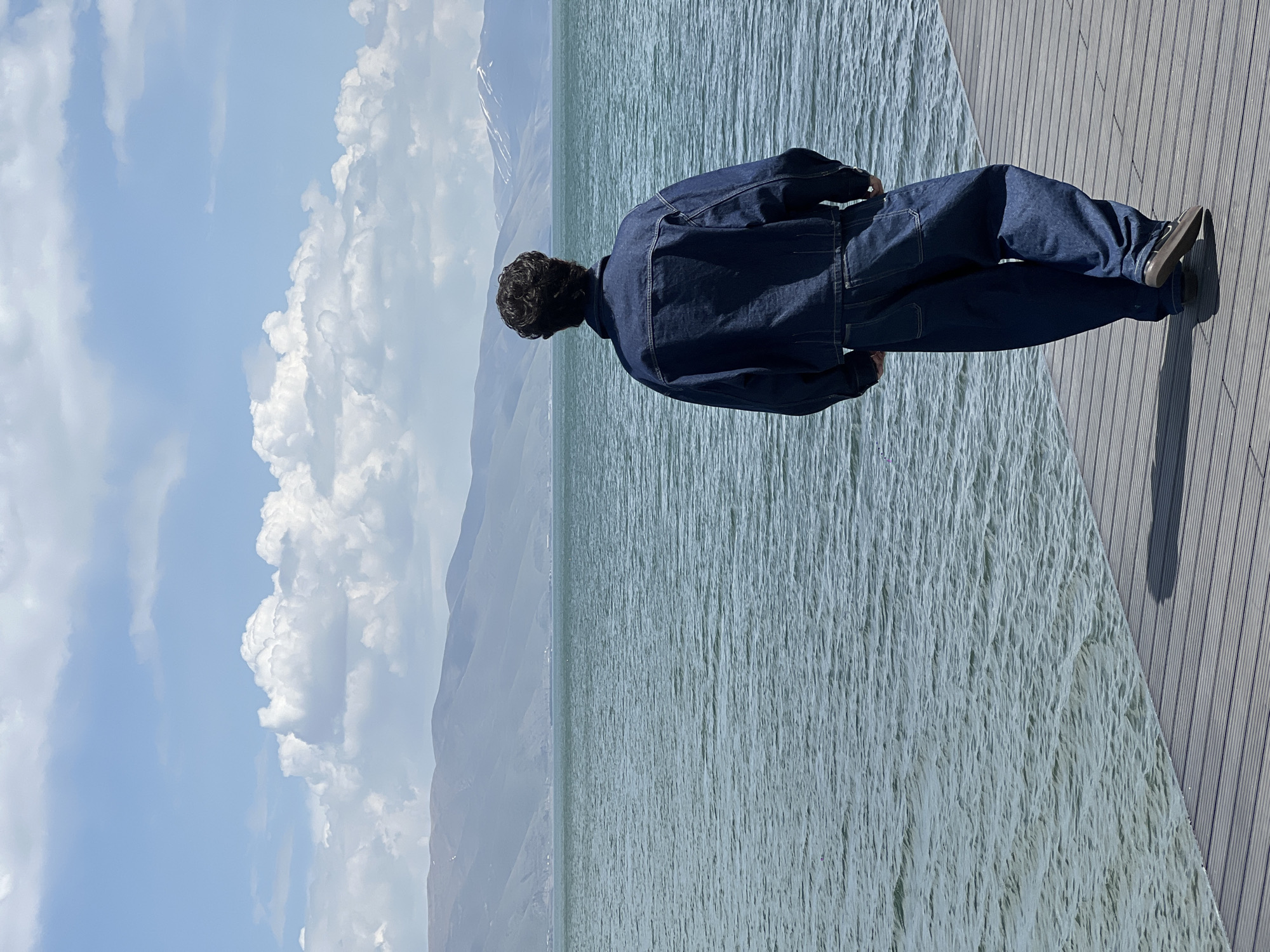A person stands on a wooden dock overlooking a body of water. The individual wears a dark blue denim jacket and matching pants, with dark shoes. Mountains are visible in the distance, partially covered with snow. The sky is partly cloudy. The location appears to be near the city of Unknown, Unknown.