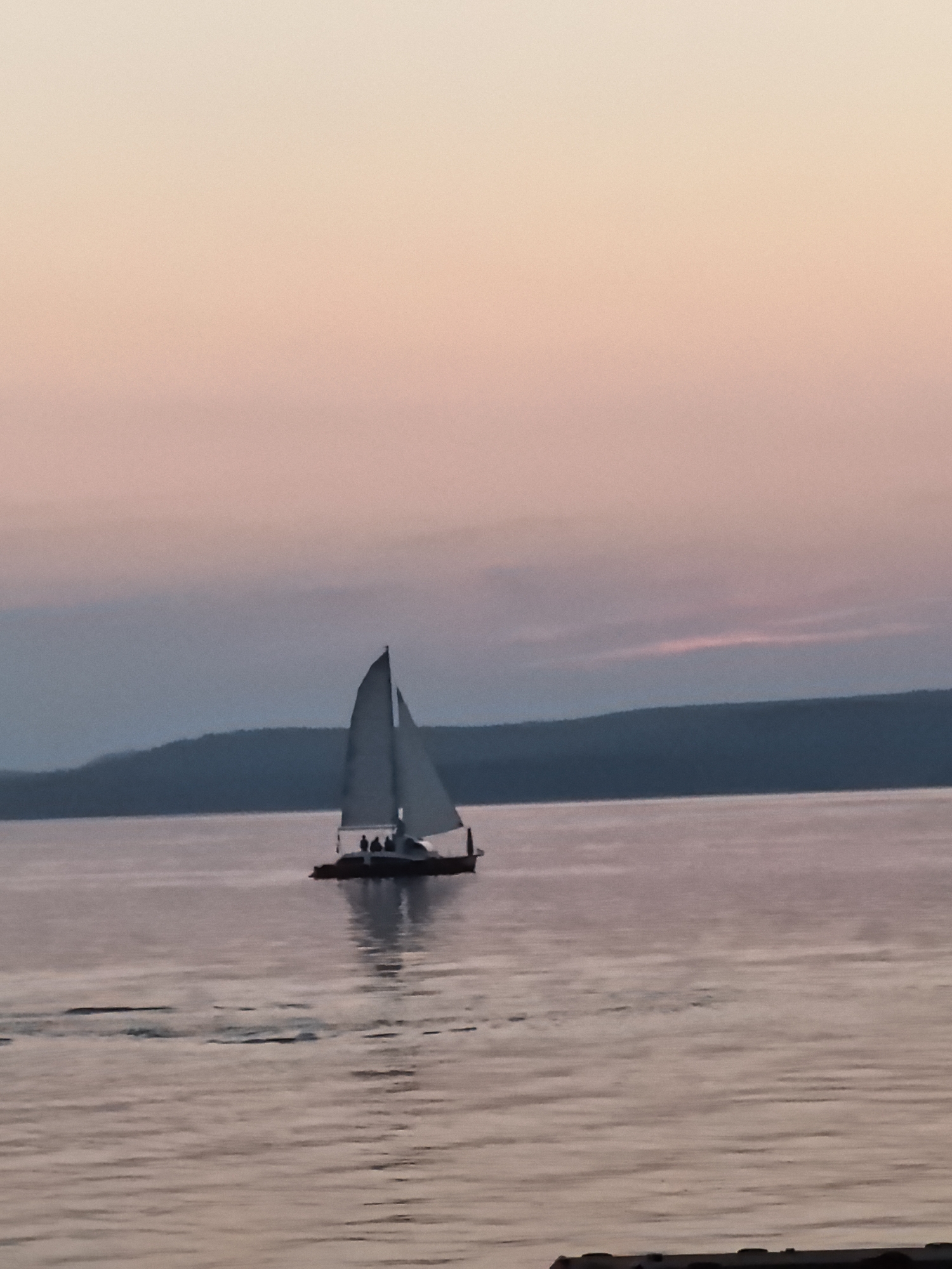 A sailboat with several people on board sails on a calm body of water at dusk. The sky is a soft gradient of pink and peach, transitioning to a muted purple and gray towards the horizon. In the background, a dark, undulating silhouette of hills or mountains is visible. The water reflects the colors of the sky and the sailboat. The scene is peaceful and serene, suggesting a quiet evening on the water. The location is the city of Osa, Russia.