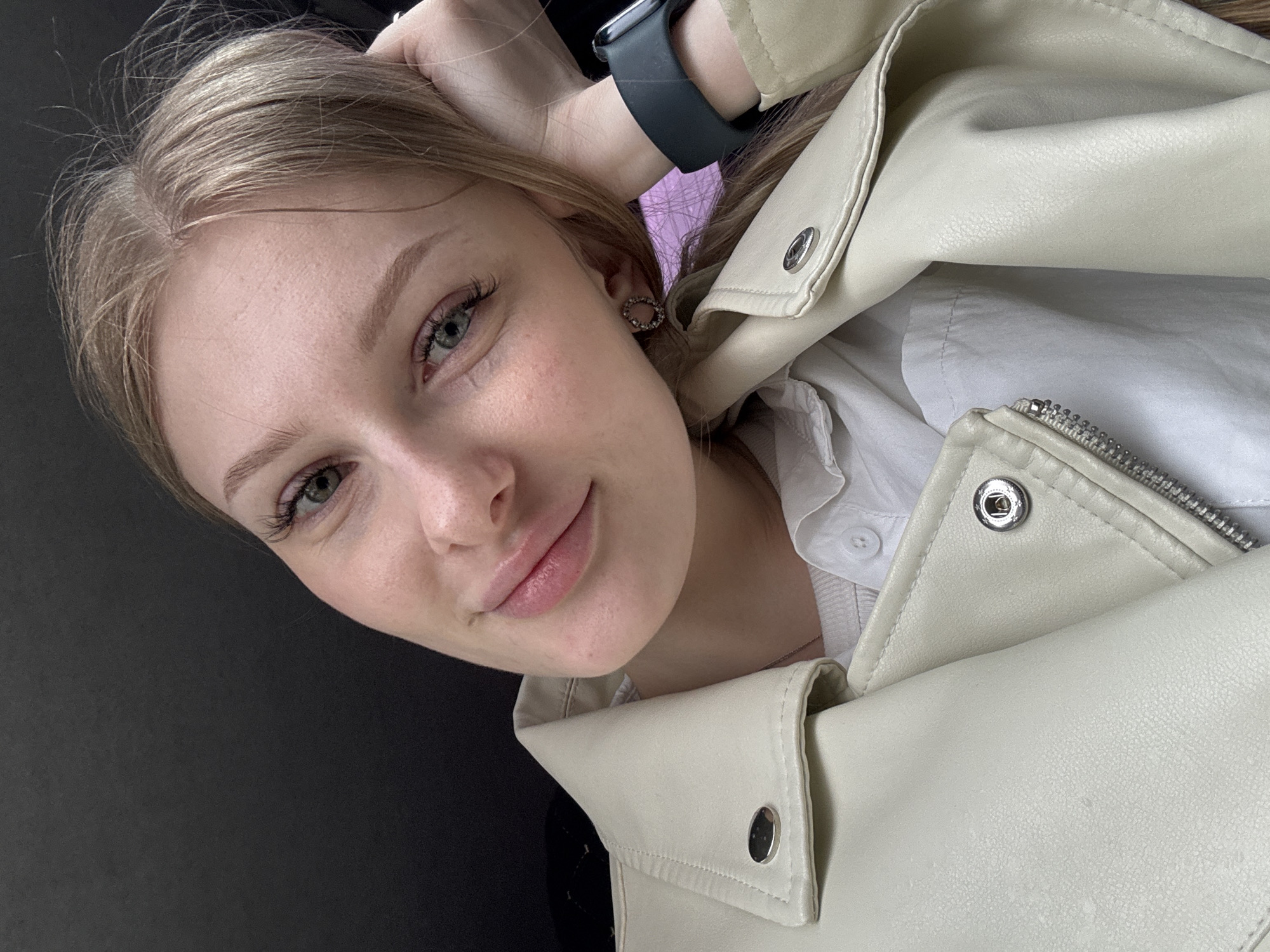 A young fair-skinned woman poses indoors, angled slightly. She wears a light cream leather jacket over a white shirt. A black smartwatch adorns her left wrist. Her hair is light brown. She is wearing circular earrings. A portion of a black surface is visible on the left side of the image. The setting is likely in Perm, Russia, based on available information.