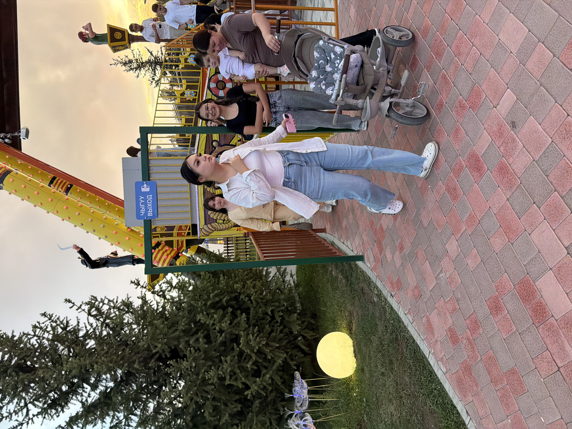 In the city of Unknown, Unknown, an outdoor amusement park scene unfolds during twilight. A paved walkway, constructed with red and grey interlocking bricks, leads the viewer's eye toward a group of people. In the foreground, a young woman with dark hair, wearing a light-colored striped shirt and blue jeans, walks with a pink smartphone in her hand. Beside her, a stroller with a patterned seat cover is partially visible.
Behind her, another young woman with glasses and dark hair, wearing a black top and grey jeans, stands by a fence. Further back, a woman in a brown shirt holds a baby wearing a white onesie. A man in a white polo shirt and another man in a white t-shirt are also present in the background, near a ride structure that features yellow, orange, and red elements, and decorative balls. The ride itself has a pirate-like theme, with figures of a pirate and a ship's wheel visible.
To the left of the pathway, a large, conical evergreen tree stands prominently. Adjacent to the tree, a bright, spherical yellow light illuminates the grassy area, with some decorative, translucent flower-like sculptures nearby. The sky above is overcast with soft, diffused light, suggesting either dawn or dusk. A blue sign with text in Cyrillic, "ВЫХОД" (Exit), is attached to a railing of the amusement ride. The overall impression is of a family outing at an amusement park as evening approaches.