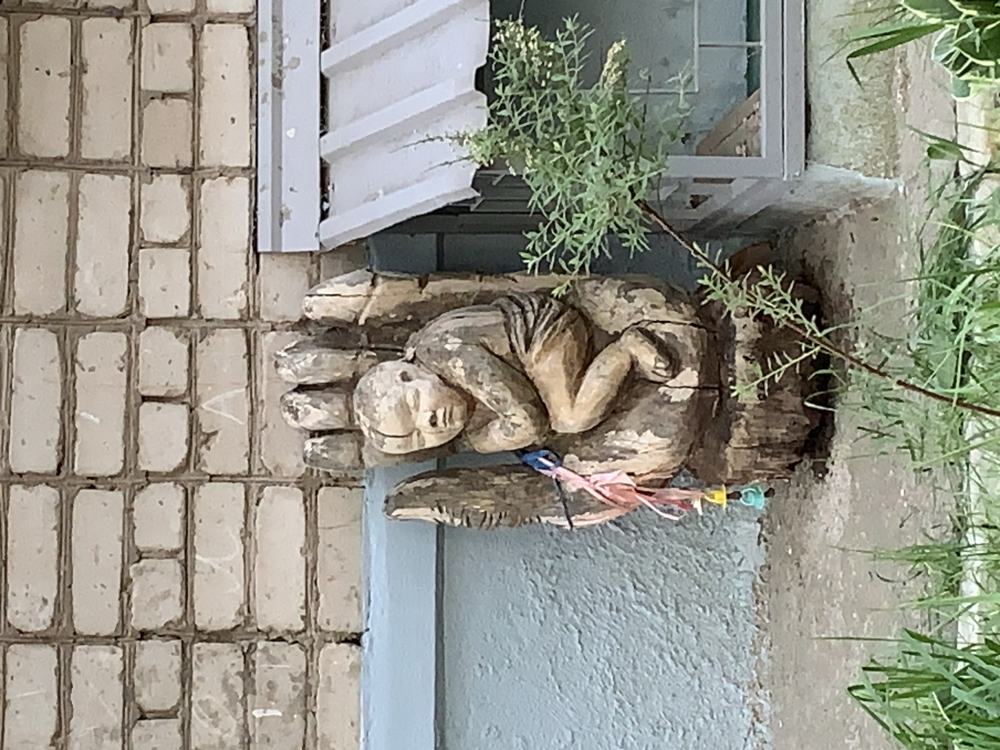 A wooden sculpture depicting a child nestled in a hand is affixed to an exterior wall in what appears to be Chaikovsky, Russia. The sculpture is located near a window with a partially open white louvered shutter. A small bush or plant is growing in the vicinity of the sculpture and window. The wall behind the sculpture is constructed of light-colored, rectangular bricks. Ribbons or fabric scraps are tied to the sculpture. The environment consists of a combination of construction materials and vegetation.