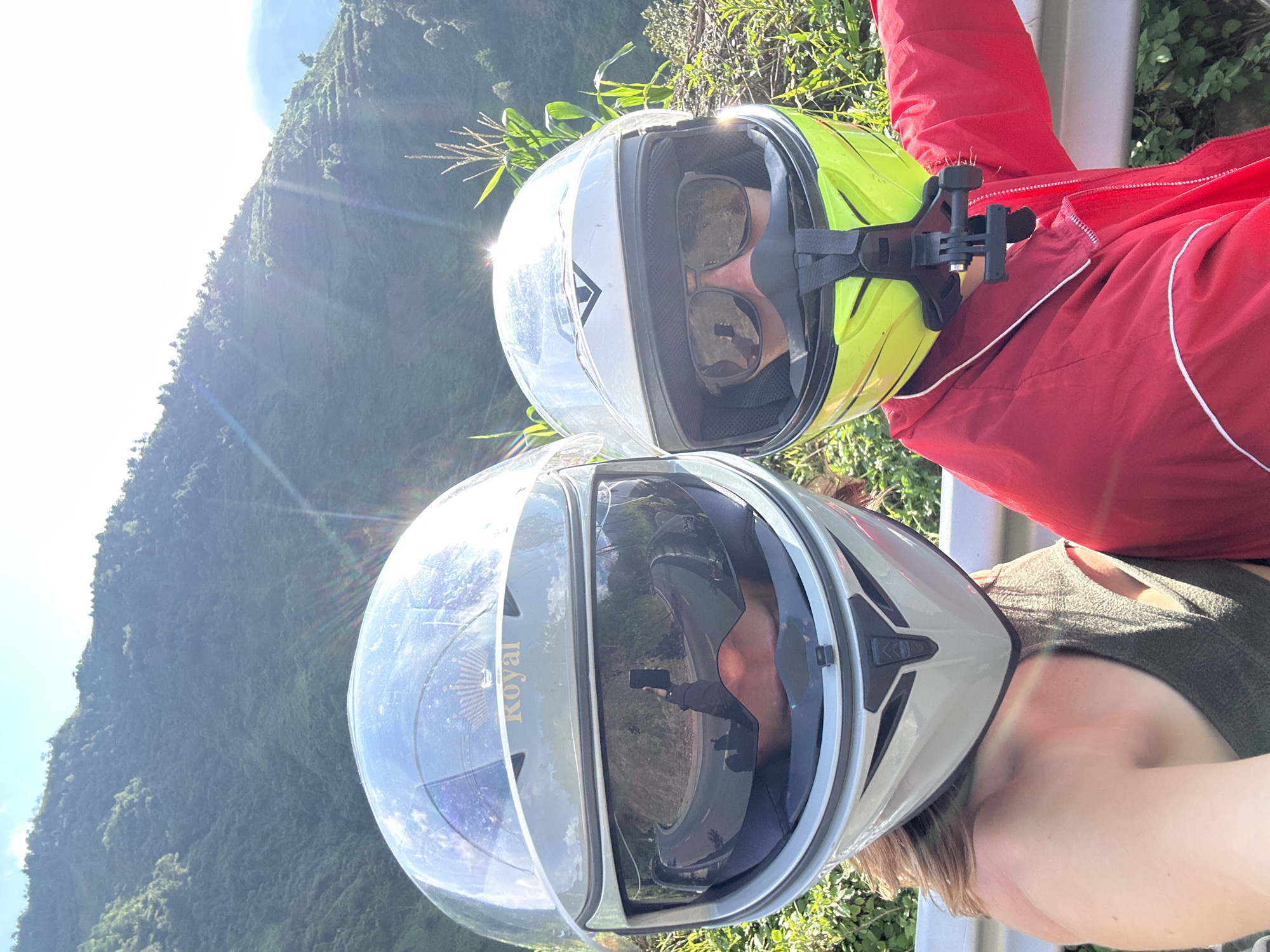 Two individuals wearing full-face motorcycle helmets are depicted in a close-up selfie taken on a sunny day in the mountainous region of Tú Lệ, Vietnam. The person on the left wears a silver "Royal" branded helmet with a reflective visor that shows a sharp curve in a paved road and the reflection of an arm holding a smartphone used to capture the image. This individual has visible light brown hair and wears a dark sleeveless top. The person on the right wears a white and neon yellow helmet with an attached GoPro chin mount, sunglasses visible under the raised visor, and a red jacket with white trim. Both subjects are positioned alongside a metal guardrail, with dense green vegetation, including corn stalks, visible in the foreground and a vast, verdant mountain range under a clear sky in the background. Sun flares are prominent, indicating direct sunlight from the upper right.