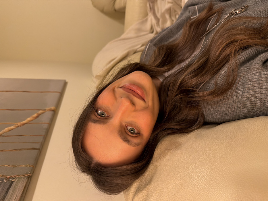 A woman with long, wavy brown hair and green eyes lies on a cream-colored leather couch. She wears a grey sweater with a silver zipper and chain. Her face is slightly tilted towards the camera, and she has a subtle smile. A painting with a gray and brown design is visible in the background, mounted on a light-colored wall. The scene is softly lit, creating a warm and inviting atmosphere.
