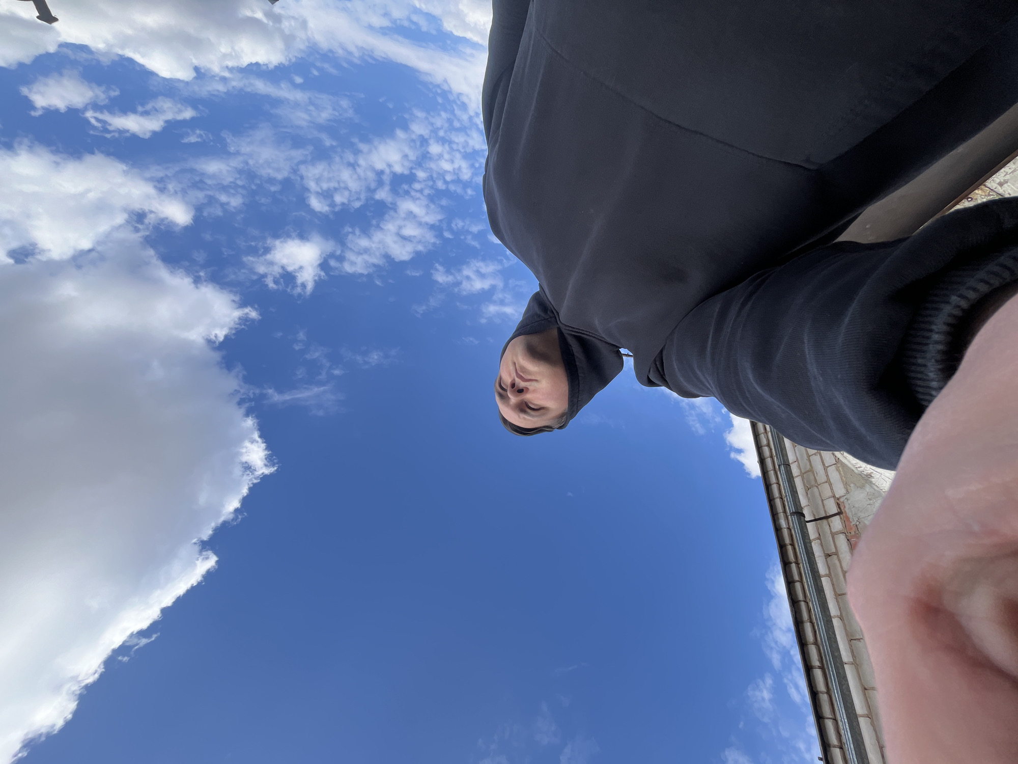 The image shows a low-angle selfie taken outdoors in Vysokovsk, Russia. A person wearing a dark hooded sweatshirt is in the frame, with the camera pointed upwards towards their face. Above the person is a bright blue sky scattered with white clouds. A section of a light-colored brick building with a dark rain gutter running along it is visible on the right side of the frame. The person appears to be holding the camera at arm's length.