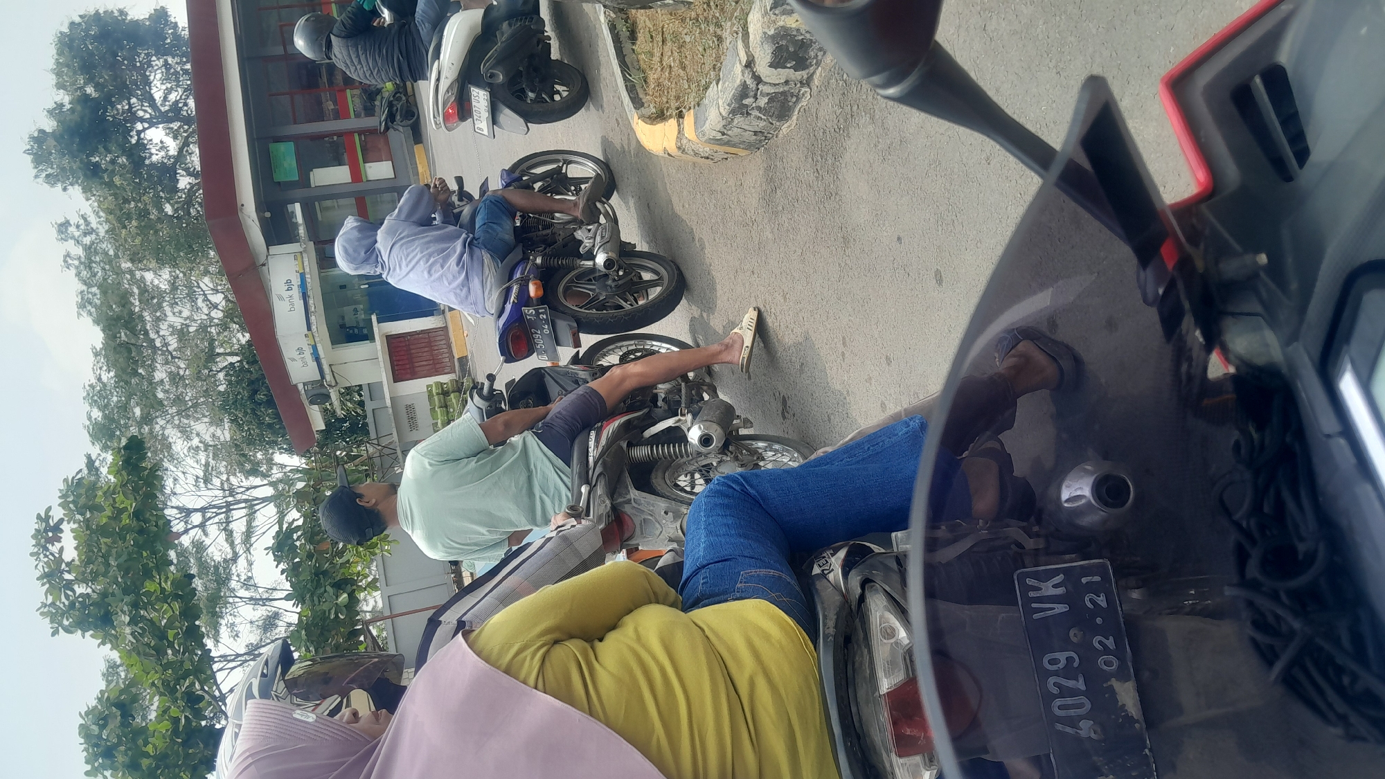 The image captures a scene at a bank in Subang, Indonesia. Several people are on motorcycles, seemingly waiting in line or at an ATM. The architecture of the bank, with its distinctive red roof and branding, is visible in the background. The motorcycles are parked on a paved road, with a curb and some greenery visible on the side. The weather appears to be overcast. One license plate on a motorcycle reads "4029 VK 02-21". The individuals are dressed in casual attire, with some wearing helmets and others not.