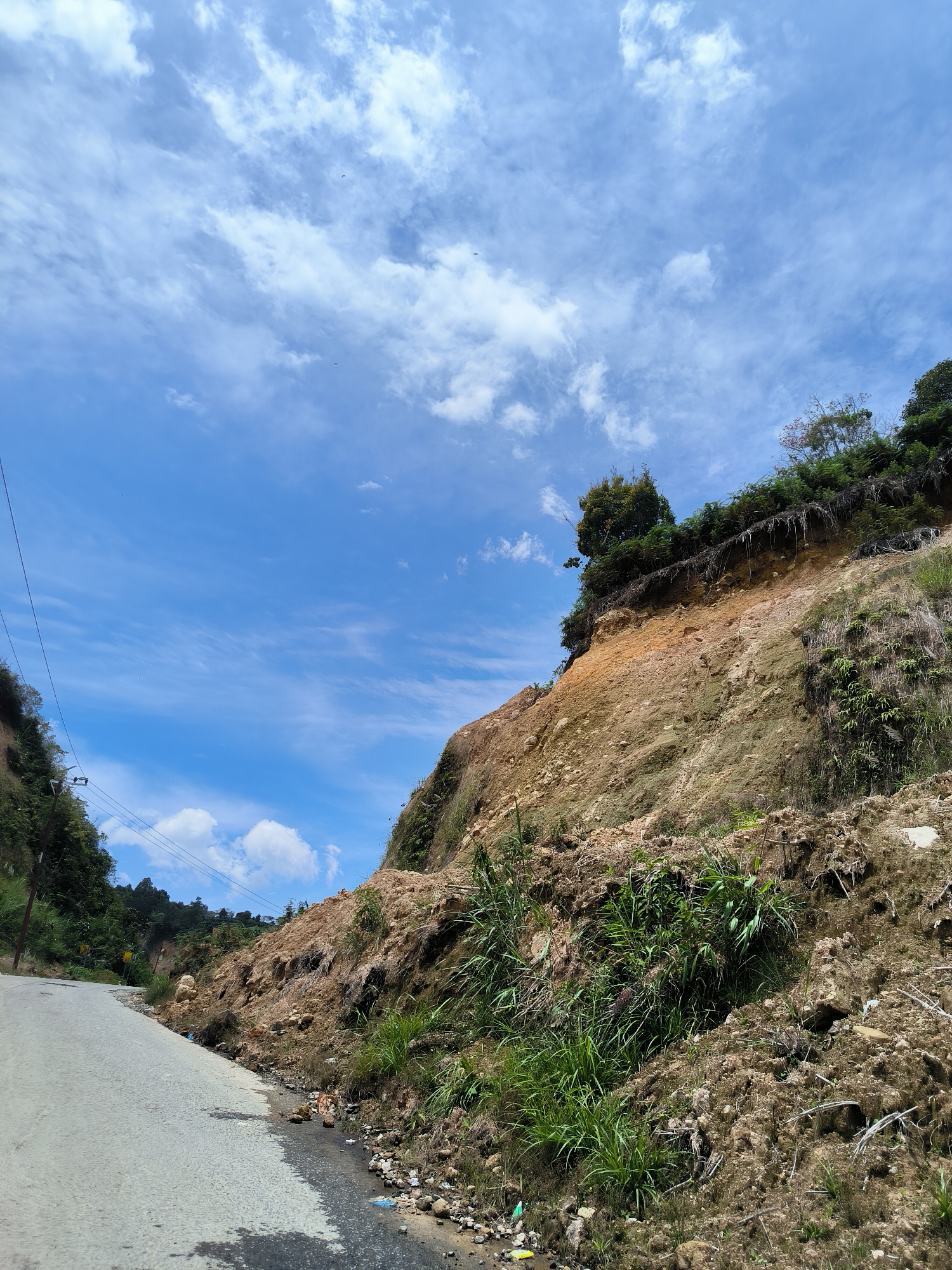 A landslide has occurred on a steep, vegetated hillside next to a paved road in Bintangpermata, Indonesia. The landslide appears to be recent, with disturbed earth and debris covering the road's edge and a small stream. The hillside is a mix of exposed soil, rocks, and patches of greenery, with trees and dense vegetation at the top. The sky is blue with scattered white clouds. Power lines are visible along the left side of the road. No people or vehicles are present.
