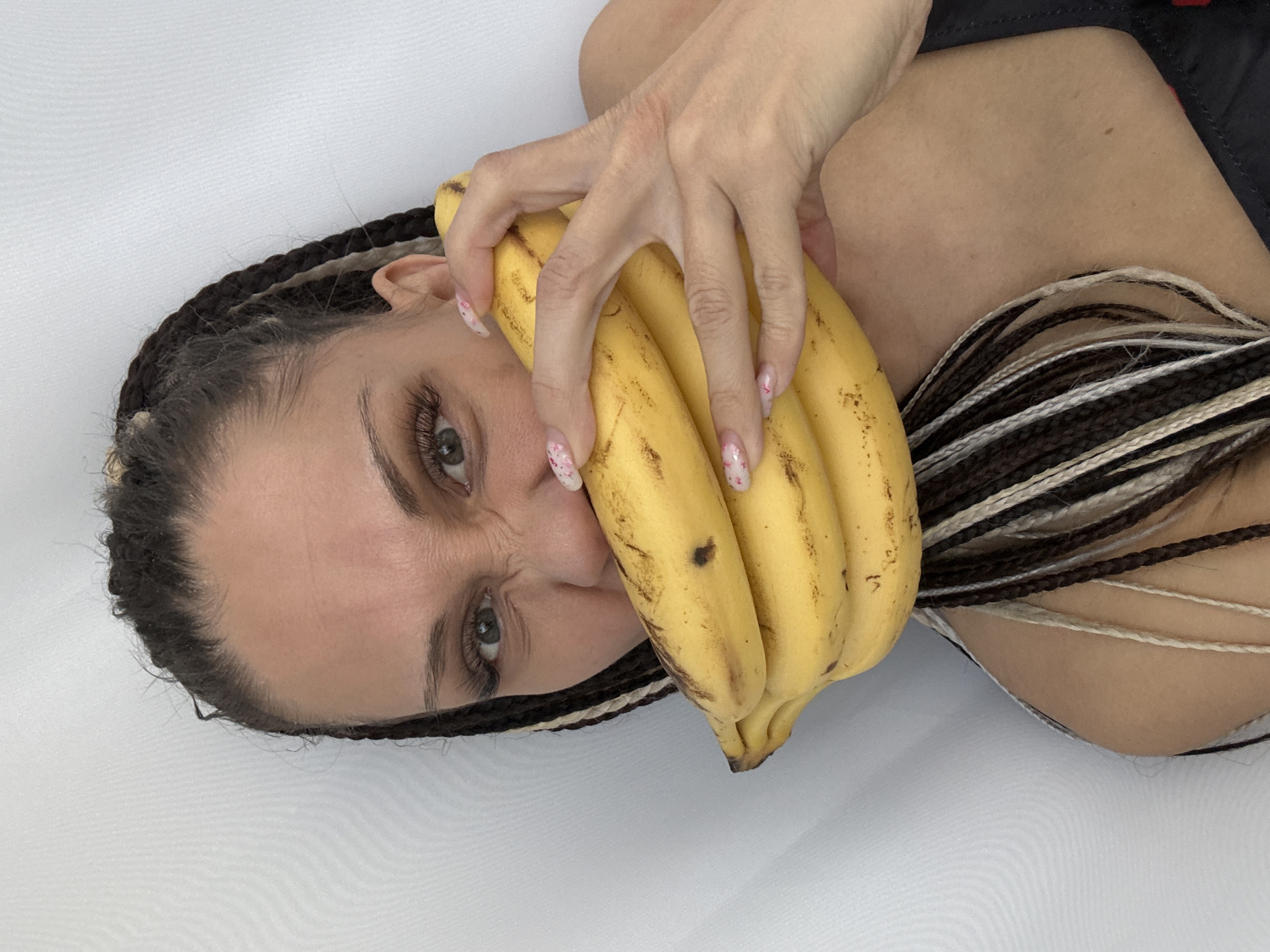A woman with braided hair is lying on a white surface. She is holding a bunch of bananas in front of her face. Her hand is resting on the bananas, with her fingers splayed. The nails on her hand are painted with a floral design. She has an intense expression on her face. The setting is unknown. Rusanovo, Russia is not evident.