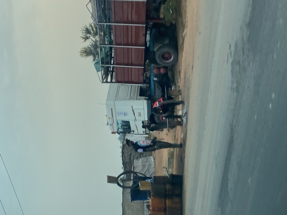 A large truck with a red cargo container on the back is parked beside a road. Two people work on the truck's tire. A man wearing what appears to be a uniform and holding a rifle stands guard near the truck. A stack of barrels and a metal hoop are near the guard. A small building is beside the road.