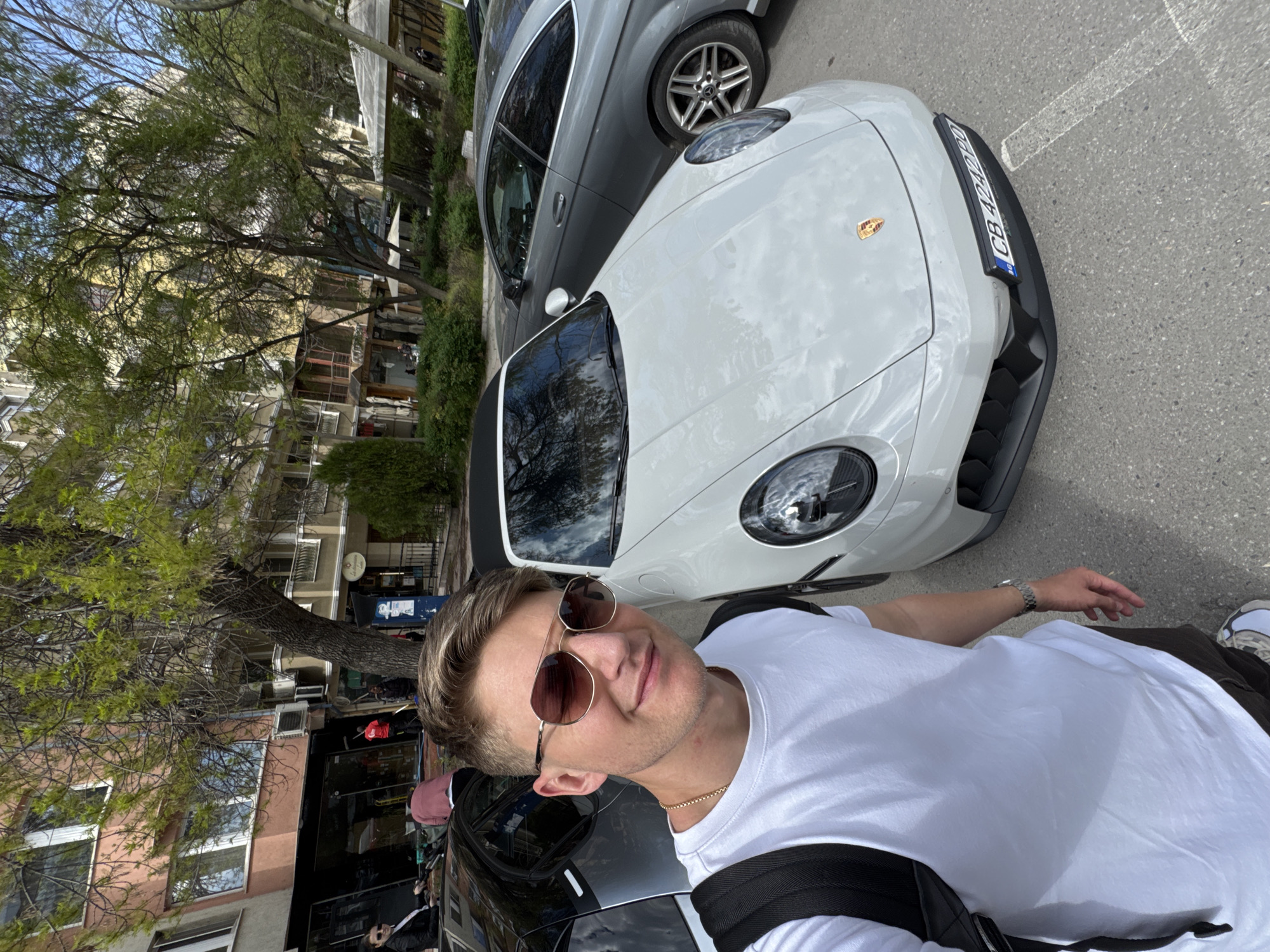 A rotated selfie-style image captures a young man posing beside a white Porsche sports car on a city street. The man, in the foreground, has short, light brown hair and wears sunglasses, a white t-shirt, and a gold chain. A dark backpack strap crosses his right shoulder, and a silver watch is on his left wrist. He has a subtle smile, looking towards the camera.
The central focus is a modern white Porsche, showcasing its sleek design, prominent front bumper, and round headlights. Its Bulgarian license plate, "CB 4242BO," is clearly visible, with "BG" (for Bulgaria) on the blue EU strip. The Porsche crest adorns the car's hood. Behind the white Porsche, a grey Mercedes-Benz sedan is parked, identifiable by its distinctive wheel design. Another dark-colored car is partially visible behind the man.
The background depicts a vibrant urban environment. Multi-story buildings of various hues, including yellow/beige and reddish-brown, line the street, featuring windows and balconies. Mature deciduous trees with fresh green foliage and bushes are scattered throughout the scene, suggesting spring or early summer. The bright daylight and clear sky indicate it is daytime, possibly midday or afternoon. Faint white markings are visible on the asphalt street. The scene evokes a casual street snapshot from Plovdiv, Bulgaria.