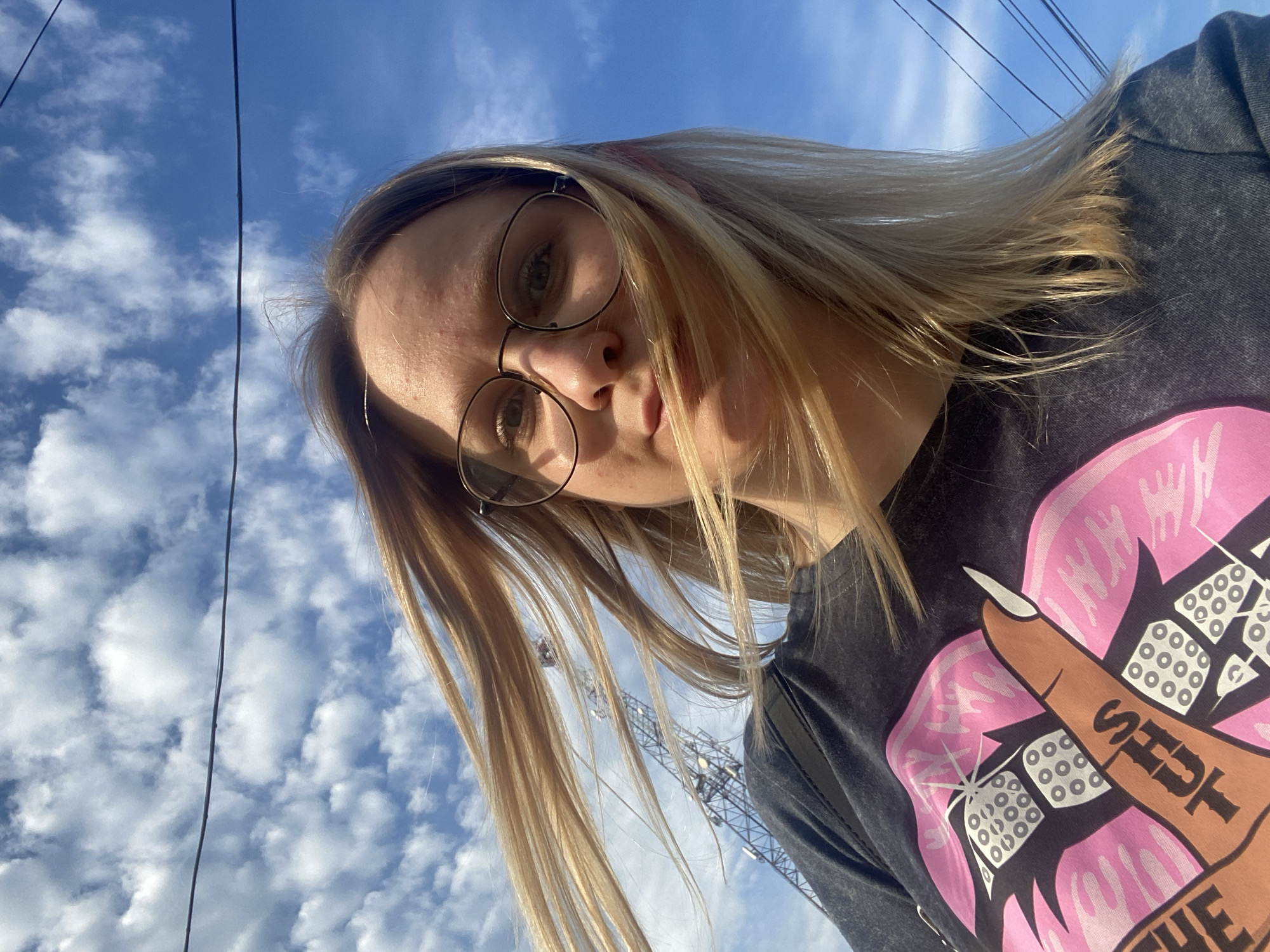 A woman with blonde hair and glasses is the subject of this eye-level selfie. The background shows a partly cloudy sky with power lines. A metal tower is visible behind the woman. The woman is wearing a dark grey t-shirt with a graphic design featuring a pink design and finger with the word "SHUT" on it. It is not possible to determine the exact location within the city of Chaikovsky, Russia, based on the image.