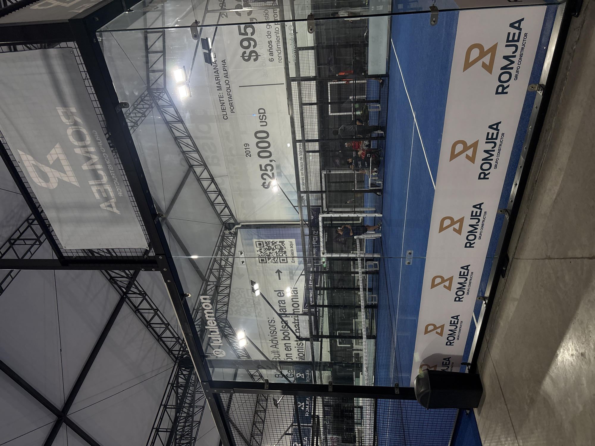 The image shows the interior of a padel tennis court in San Bernardino Tlaxcalancingo, Mexico. The court has glass walls adorned with advertising. "ROMJEA, Grupo Constructor" is visible on a banner near the base of the wall. Above that is advertising that says "@lululemon" and "BlBull Advisors: Geón en bolsa para el...ionist natrimoniai." On another wall, advertising states "CLIENTE: MARIANA L. PORTAFOLIO ALPHA," with financial details "$25,000 USD," and "$95." Players are seen on the blue court. The structure of the building is evident in the ceiling framework. A QR code with the text "Empieza aqui" is on one of the walls. There's also a sign that says "6 anos de gestio rendimento prome..."