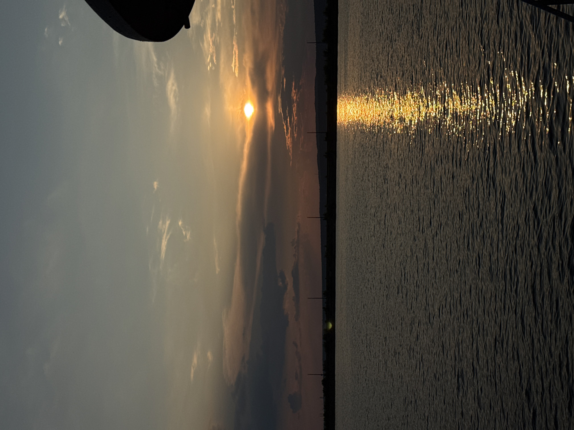 The image depicts a sunset over the Kama River in Chaikovsky, Russia. The sun, a bright orb, is partially obscured by clouds, casting a golden reflection on the water's surface. The foreground is dominated by the dark, rippling water. In the background, a silhouetted treeline and scattered utility poles mark the distant shore. The sky exhibits a gradient of colors, from pale blue at the top to warmer oranges and reds near the horizon, with wispy clouds scattered throughout. A dark, curved object, possibly part of a structure or vessel, is visible in the upper right corner of the frame.