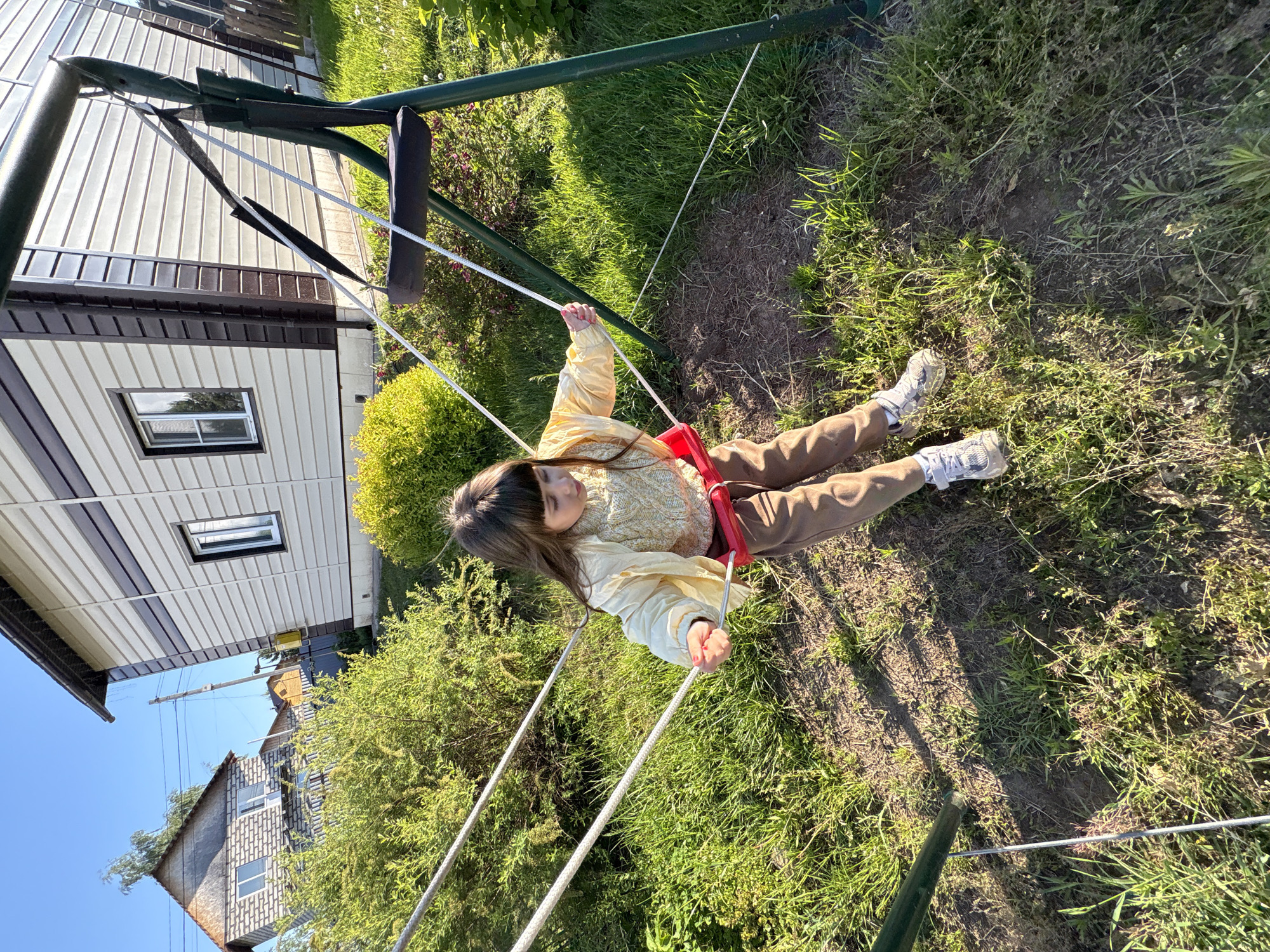 A young girl is captured mid-swing on an outdoor swing set in Chaikovsky, Russia, during what appears to be a bright, sunny day. She has long, dark hair and is wearing a light yellow long-sleeved top, brown pants, and white sneakers. Seated on a red swing seat, she holds onto the white ropes attached to a dark green metal frame, with a calm and relaxed expression.
The scene is set in a green, natural environment, likely a backyard or garden. The ground beneath the swing is a mix of vibrant green grass and patches of bare earth or dried vegetation. Strong sunlight creates distinct shadows across the ground. Various green bushes are scattered around the swing structure and in the background.
In the background, two houses are visible. One house to the left features light-colored horizontal siding with dark brown trim and a single square window. Further in the distance and slightly to the right, another house, possibly built with brick or stone, can be seen against a clear blue sky, along with utility poles and wires. There is no visible text in the image. The overall atmosphere suggests a serene and leisurely outdoor moment.