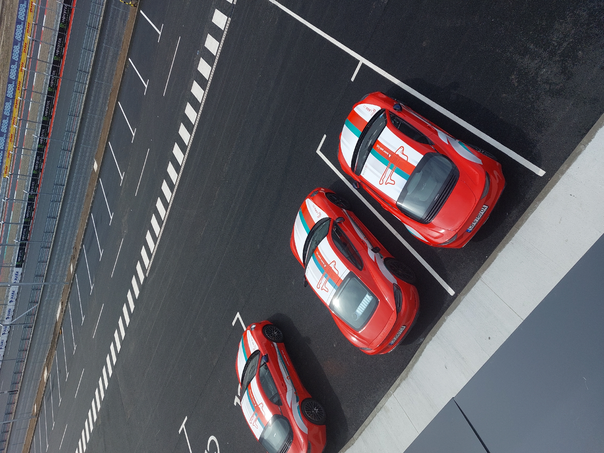This aerial view captures a parking lot, possibly near a racetrack in an unknown city, containing three red cars parked at an angle. The cars feature a white racing stripe and a logo consisting of a stylized outline of a person with a race track shape running through the figure.
The parking lot itself has standard white lines defining the parking spaces. In the upper left of the frame, there is a barrier wall with advertisements from DHL and Monster Energy. The ground is black asphalt, and the overall image suggests an organized racing or driving event.