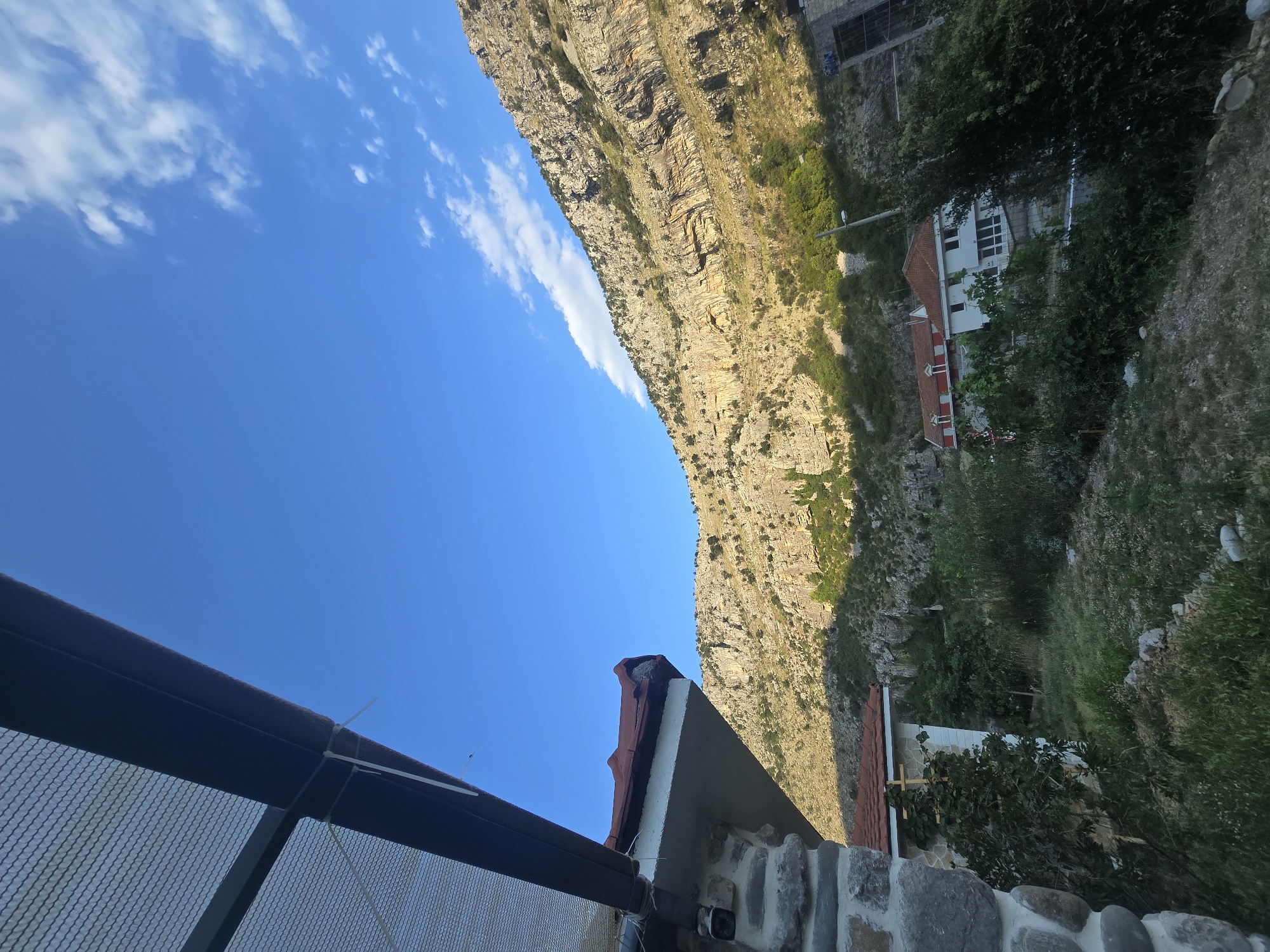 The image depicts a view from a sheltered area looking out towards a mountainous landscape with some buildings nestled at its base. In the foreground, the edge of a structure with a red-tiled roof and stone detailing is visible on the left, partially obscuring the scene. A metal mesh-like material is also present at the top left.
The main focus of the image is a large, rocky mountain face that rises steeply from the valley. The mountain exhibits layers of rock and is sparsely covered with vegetation, primarily small trees and shrubs. Sunlight illuminates the mountain, casting shadows and highlighting its textured surface.
Below the mountain, several buildings with red-tiled roofs are visible. One building appears to be white with multiple windows, and another is partly red. These structures are situated amidst greenery, including trees and bushes. The environment appears to be a rural or semi-rural setting, possibly a village or a residential area situated in a valley near a mountain range. The sky is a clear, bright blue with a few wispy white clouds.
The scene suggests a location in Podgorica, Montenegro, characterized by its proximity to mountainous terrain and the Mediterranean architectural style of the buildings.