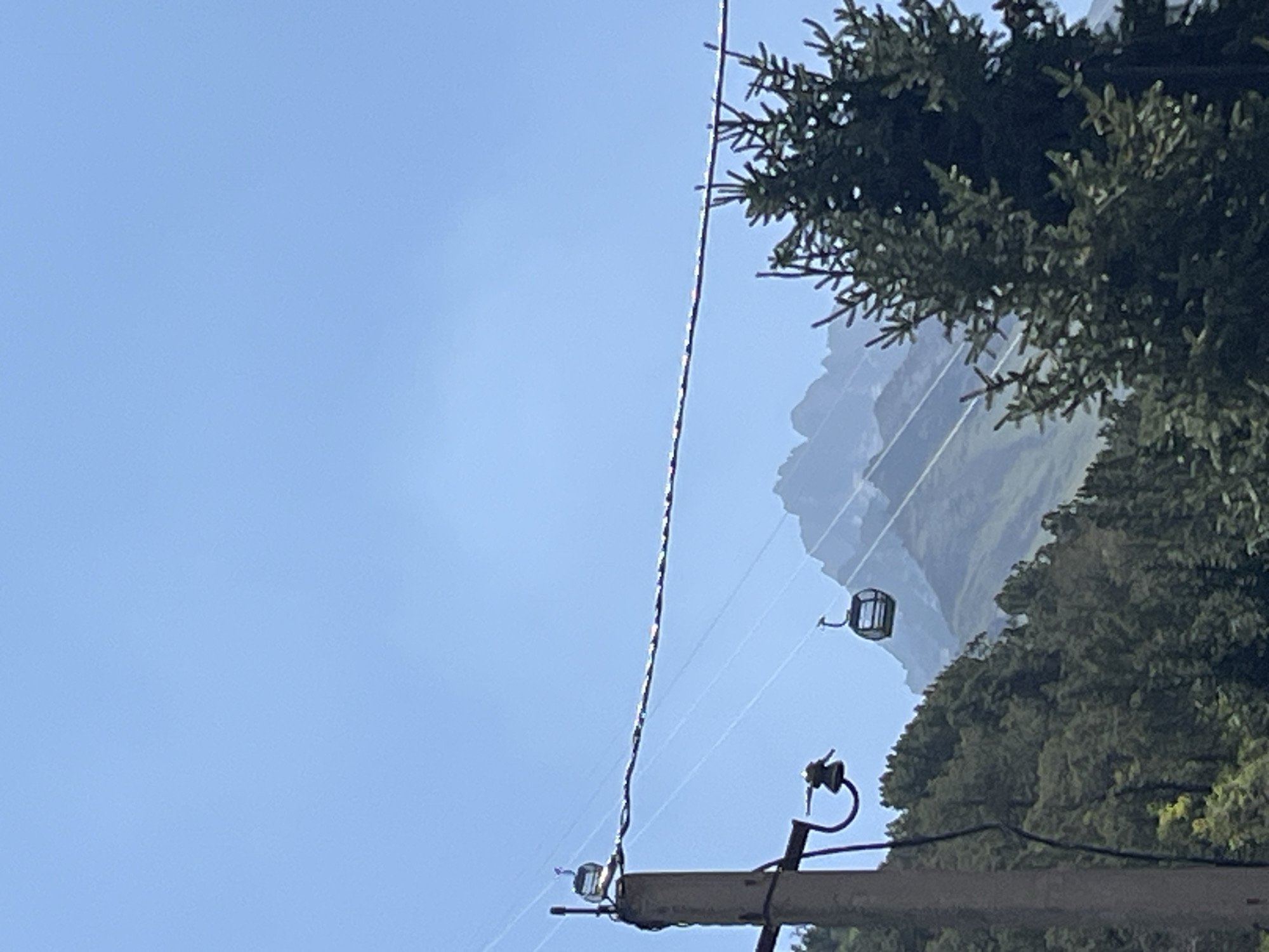 The image shows a clear blue sky over a mountainous landscape, likely in the vicinity of Kashka Suu, Kyrgyzstan. In the foreground, a utility pole with wires and what appears to be an insulator is visible on the left. Two cable cars are suspended on wires spanning across the frame. One cable car is positioned higher up, closer to the utility pole and the sky. The other cable car is lower and further in the distance. A dense evergreen forest covers the lower slopes of the mountains. In the background, jagged mountain peaks are visible, partially obscured by atmospheric haze. A large evergreen tree dominates the right side of the frame, obscuring some of the view of the mountains and cable cars. The scene appears to be captured during daylight hours.