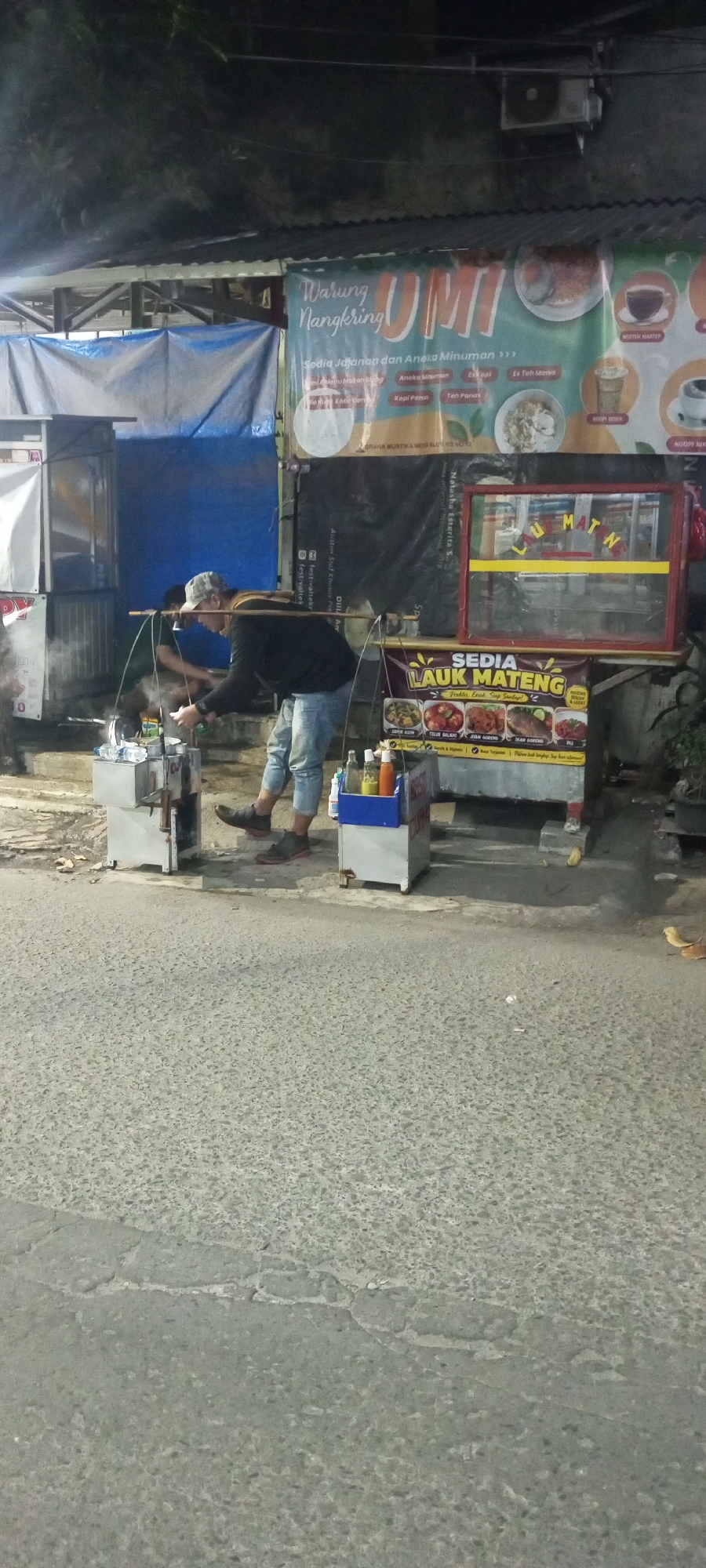 The image depicts a street food vendor in Setu, Indonesia, at dusk. A man, wearing a grey baseball cap, a dark long-sleeved shirt, and light blue jeans, is bent over a portable cooking station. He is holding a ladle and appears to be preparing food. The station features a metal frame with a blue tarp overhead and various containers, including what looks like a water cooler. To his right, another vendor is visible, also bent over a similar setup.
Behind the vendors, a banner advertising "Warung Nangkring UMI" is prominently displayed. It lists various food and drink items like "Anaka Minuman" (various drinks), "Mi Goreng" (fried noodles), "Kopi Panas" (hot coffee), and "Es Teh Manis" (sweet iced tea). Another banner below reads "SEDIA LAUK MATENG" (Ready-to-eat side dishes) and showcases images of various Indonesian dishes. A red-framed display case is also visible above this banner.
The scene is set on a paved street with some debris scattered on the ground. The lighting suggests it is evening or night, with artificial lights illuminating the food stall area. The overall environment is typical of a bustling street food scene in Indonesia.