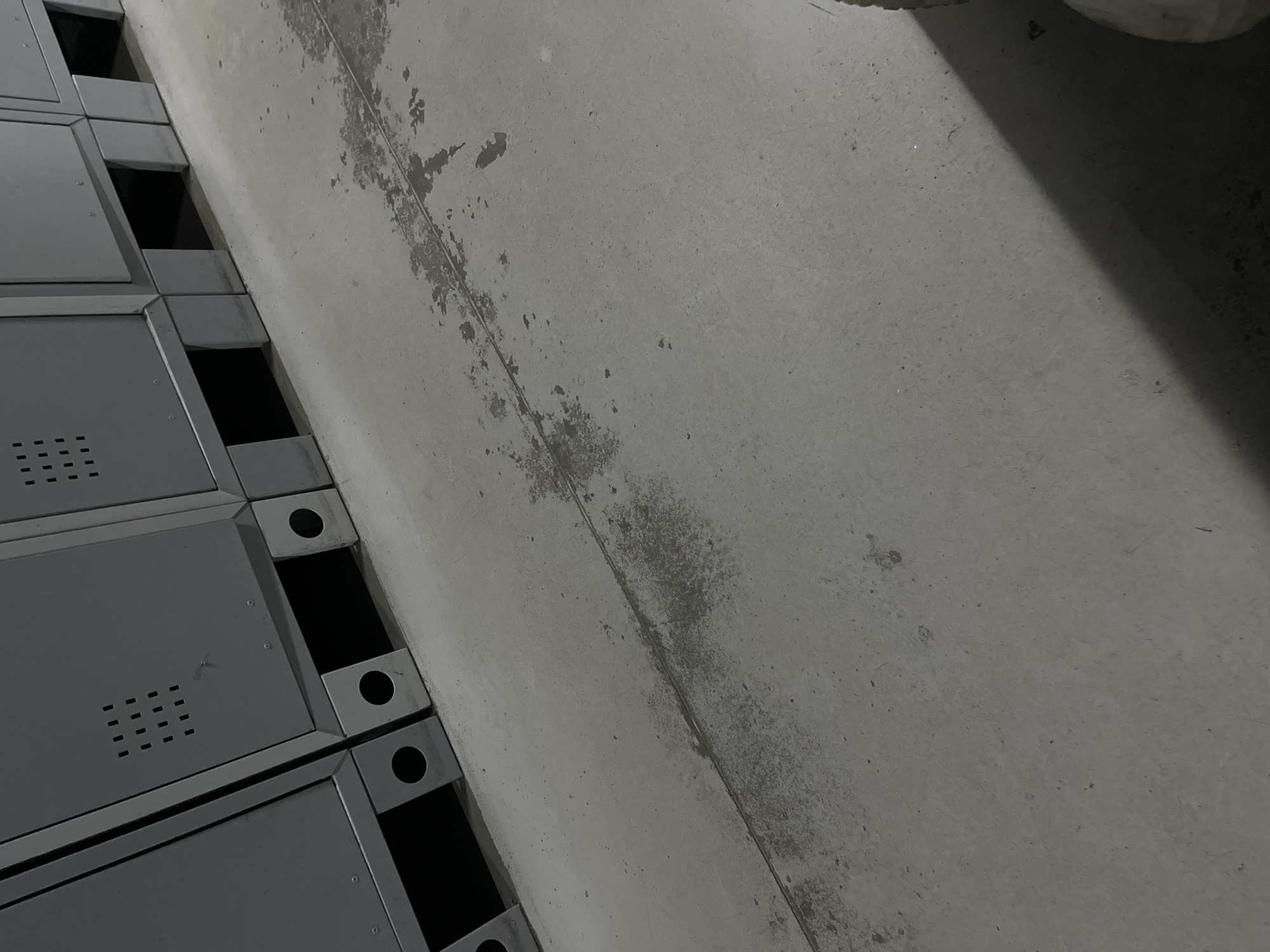 The image shows a row of gray metal lockers in what appears to be an indoor, utilitarian space. The lockers are elevated off the concrete floor by small metal risers, creating openings underneath. Some of the lockers have ventilation holes arranged in a grid pattern. The concrete floor has scuff marks and discoloration, suggesting wear and tear. A shadow is cast across the floor from the right side of the image. No people or specific landmarks are visible to determine the precise location, and therefore, the city of Kyiv, Ukraine cannot be confirmed from the visual evidence.