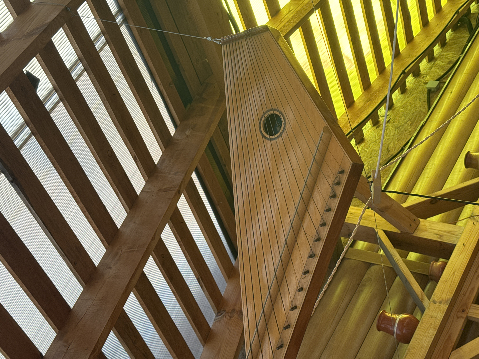 The image shows the interior of a structure, possibly a music venue or cultural center, located in Kladovitsy, Russia. A light-colored wooden harp-like instrument with multiple strings hangs suspended from the wooden framework of the ceiling. The ceiling's framework is a complex arrangement of beams and rafters, with translucent panels between some of the wooden elements, allowing daylight to filter through. Additional wooden structures and possibly ceramic resonators are visible in the background.