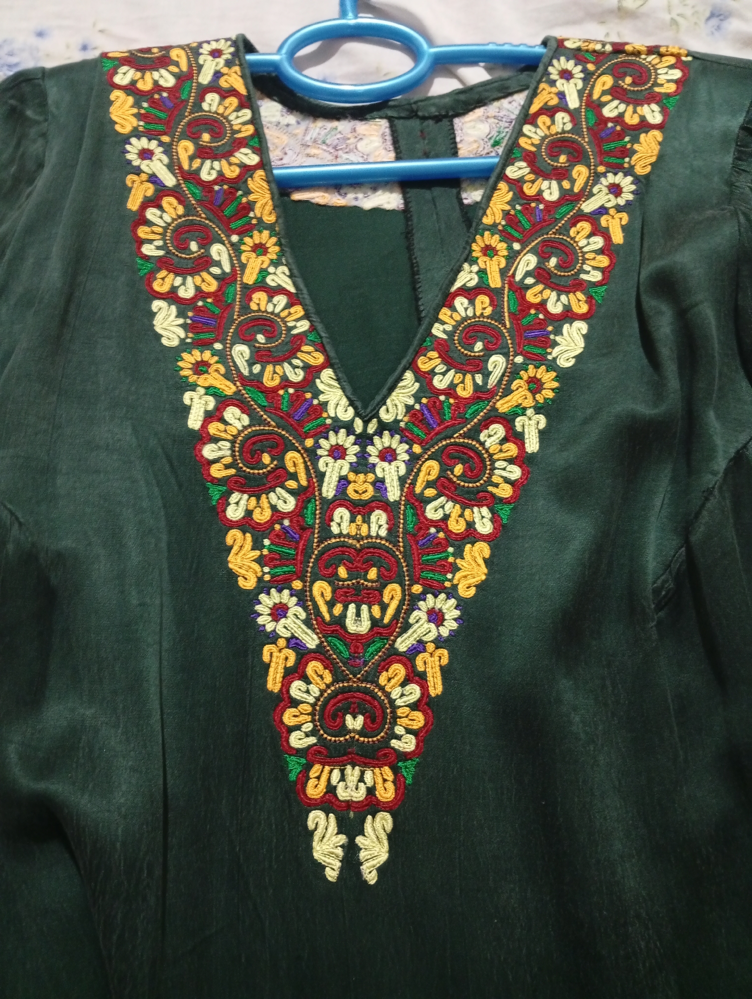 The image displays a close-up view of the neckline of a dark green garment, possibly a kurti or dress. The garment features intricate V-shaped embroidery around the neckline, extending down towards the chest. The embroidery is a vibrant mix of colors including red, yellow, white, green, and purple, forming floral and paisley motifs with swirling patterns. The fabric of the garment appears to be a smooth, slightly textured material. A blue hanger is visible at the top of the image, supporting the garment. The background is out of focus but suggests a lightly patterned fabric, perhaps a bedsheet or tablecloth. There is no indication of any specific location in the city of Seýdi, Turkmenistan.