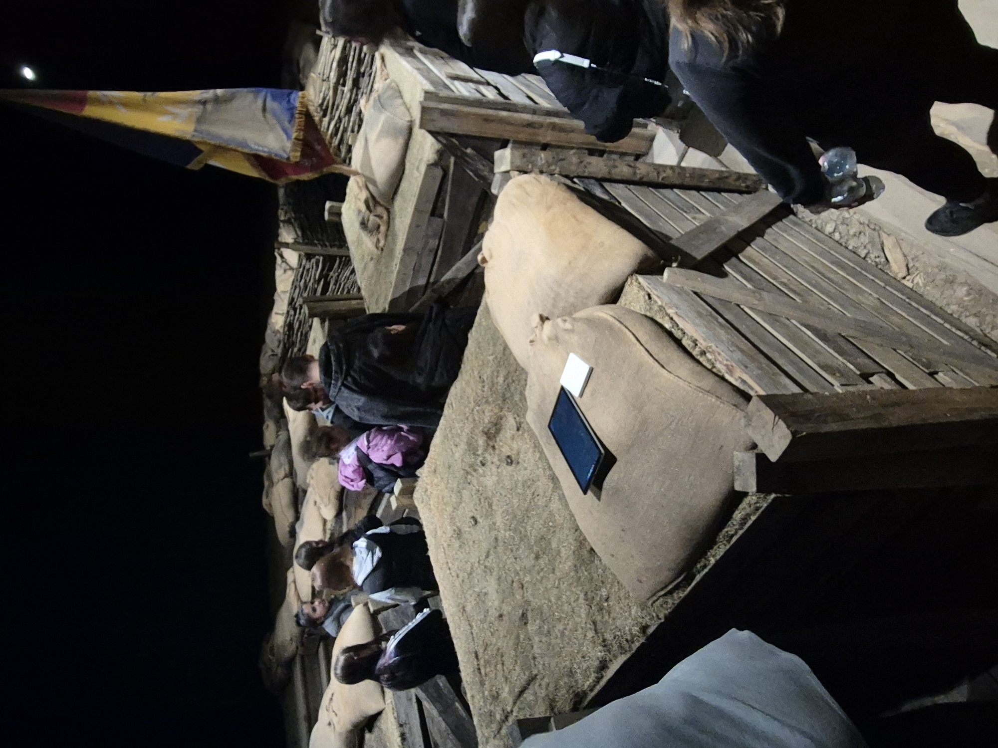 The image shows a group of people resting at night on a structure that appears to be a stone and wood building. A yellow and blue flag with a red border hangs near the structure. The setting is dark with a bright object, possibly the moon, visible in the upper left corner. People are either sitting or lying down on surfaces covered with woven mats and what seems to be stuffed cushions. One person is wearing pink while another wears a black outfit. A cell phone rests on one of the cushions. A person is carrying a water bottle. The location may be Shumentsi, Bulgaria, inferred from the prompt, but without further visual context, this cannot be definitively confirmed. The composition suggests a casual or makeshift resting area.
