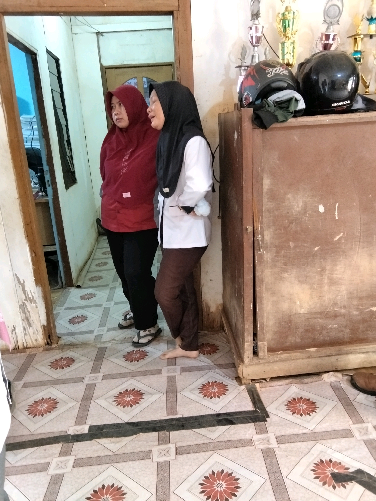 Two women are standing in a doorway. The woman on the left wears a red hijab and a red top. The woman on the right wears a black hijab, a white shirt, and brown pants. Both women appear to be speaking to each other. Behind them, a wooden cabinet holds two helmets and several trophies. The floor is tiled with a floral pattern. The room appears to be indoors, possibly in a home or office in an unknown city in Indonesia.
