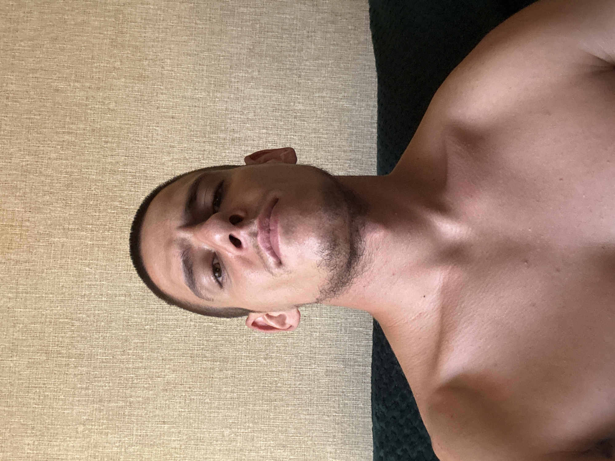A shirtless man with a shaved head and a light stubble is looking directly at the camera. He is positioned in the foreground with his chest and shoulders visible. Behind him is a textured beige wall and a dark green surface, possibly a sofa. The lighting is natural and illuminates his skin, highlighting sweat or oil. The image appears to be a selfie taken indoors. There are no specific identifiable landmarks or contextual clues to suggest a location in Sofia, Bulgaria.