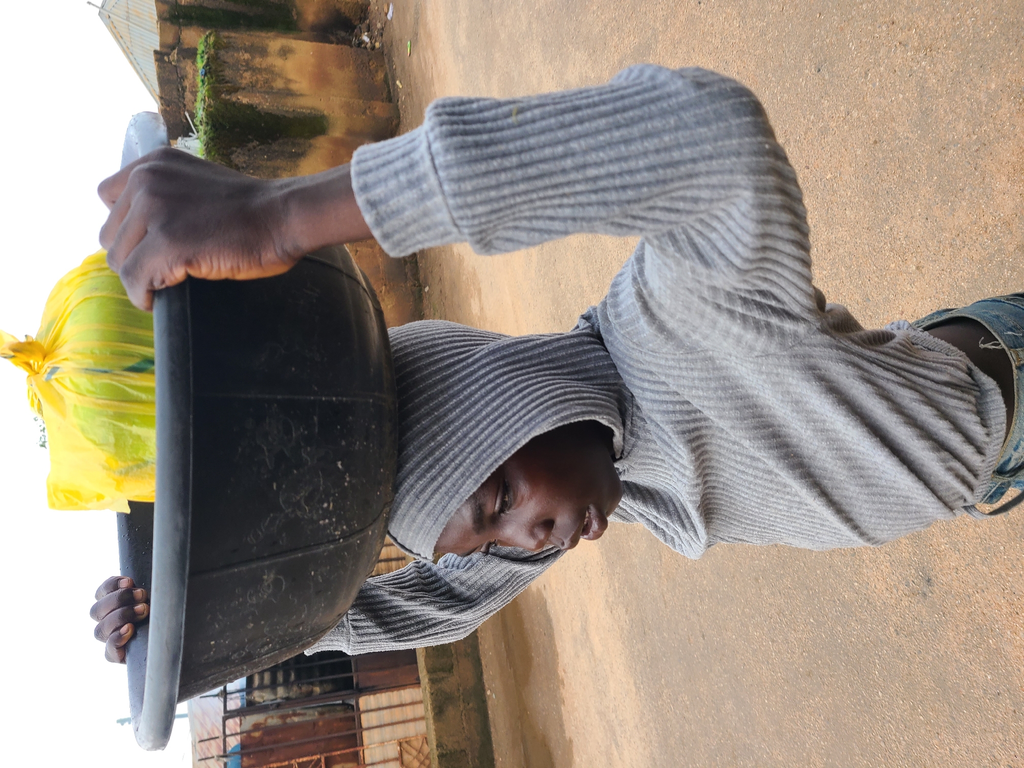 A young person, possibly a girl, is captured in a medium shot from the chest up, walking outdoors. They are wearing a gray, ribbed, hooded sweatshirt and blue jeans. The person is balancing a large, black basin filled with a bright yellow plastic bag on their head, using both hands to steady it. The background suggests an urban environment in Jos, Nigeria, with a rough, dirt road, a textured, weathered wall, and a corrugated metal roof visible. The lighting indicates it is daytime.