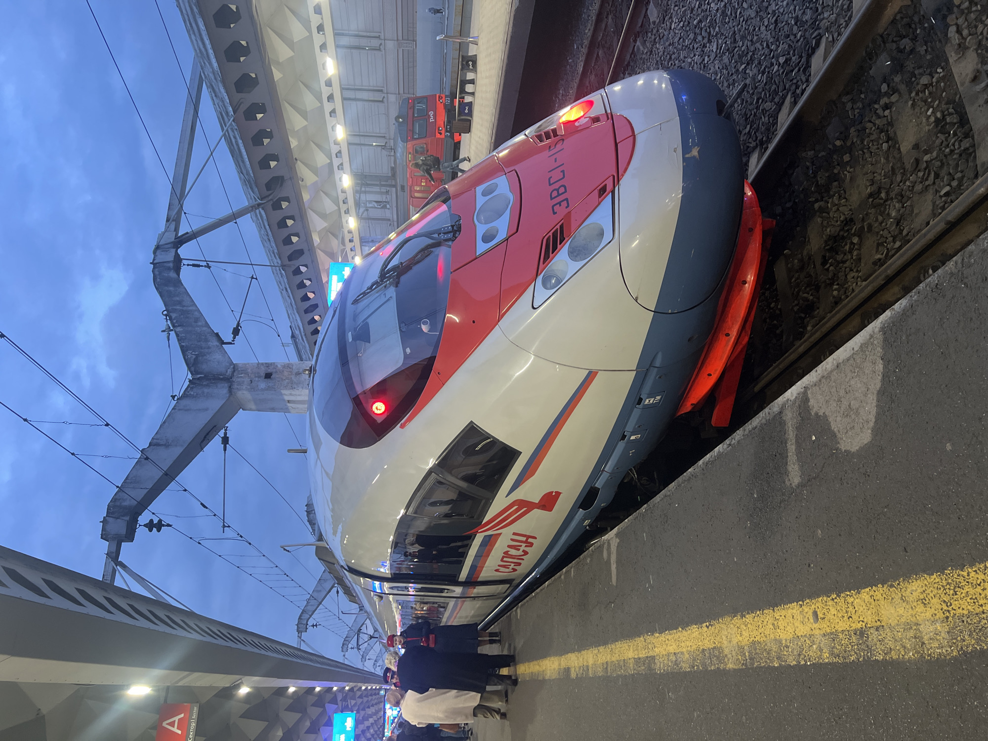 A passenger train is at the platform in a station, most likely in Saint Petersburg, Russia. The train is a Sapsan high-speed train, white with red and blue trim, and the name "САПСАН" (Sapsan) is visible on the side. Some passengers are standing near the train. The platform has a yellow safety line and a sign with the letter "A". Overhead power lines and station infrastructure are visible. In the background, another red passenger train stands on the neighboring platform. The sky above appears to be in the evening or dusk.