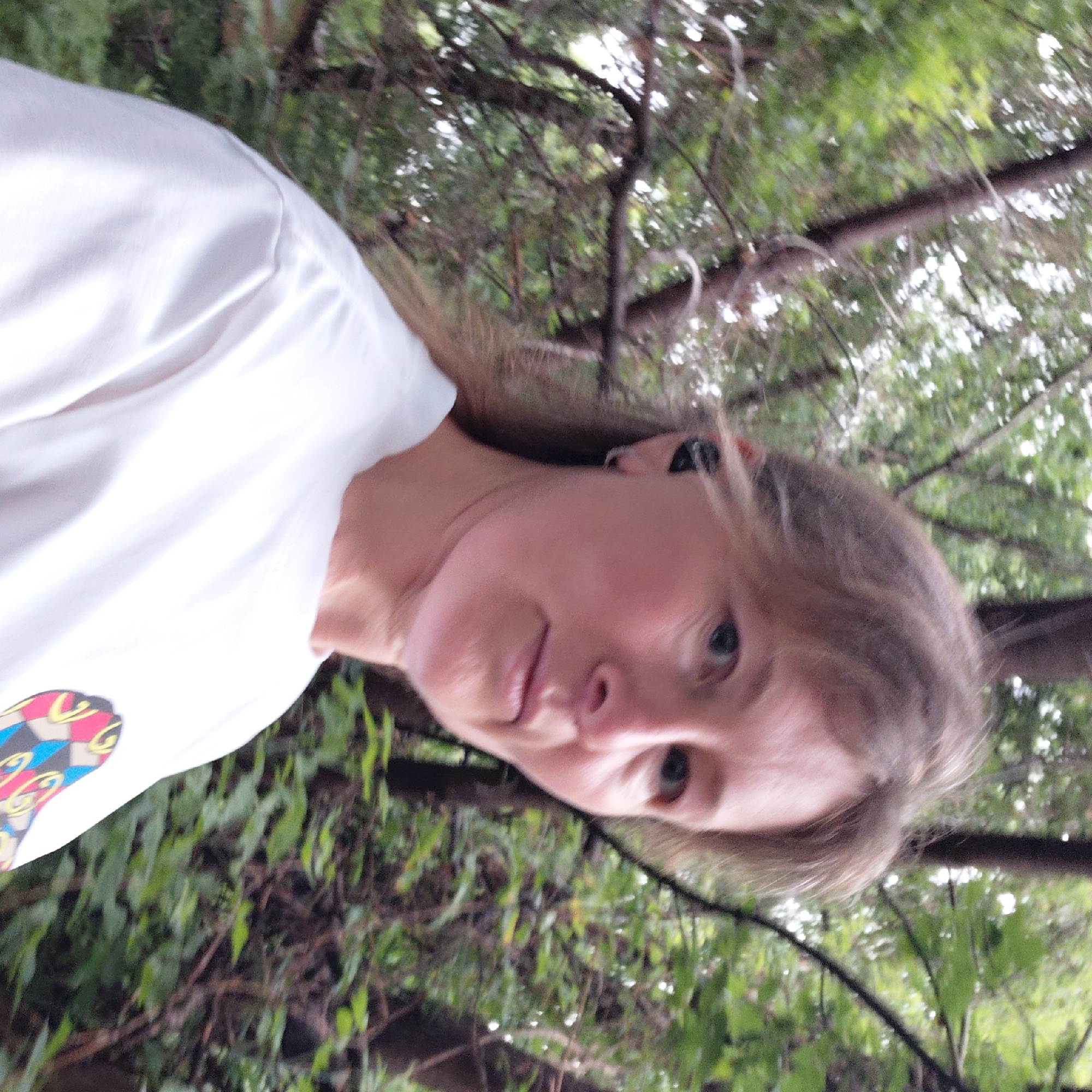 The image shows a close-up, head-and-shoulders shot of a woman with blonde hair wearing a white t-shirt. She is outdoors, with blurred trees and foliage in the background, suggesting a natural setting. She is wearing black earbuds. The woman's face is in profile, with her gaze directed slightly off-camera. The lighting suggests an overcast day or shade from the trees. The location is not discernible from the image.