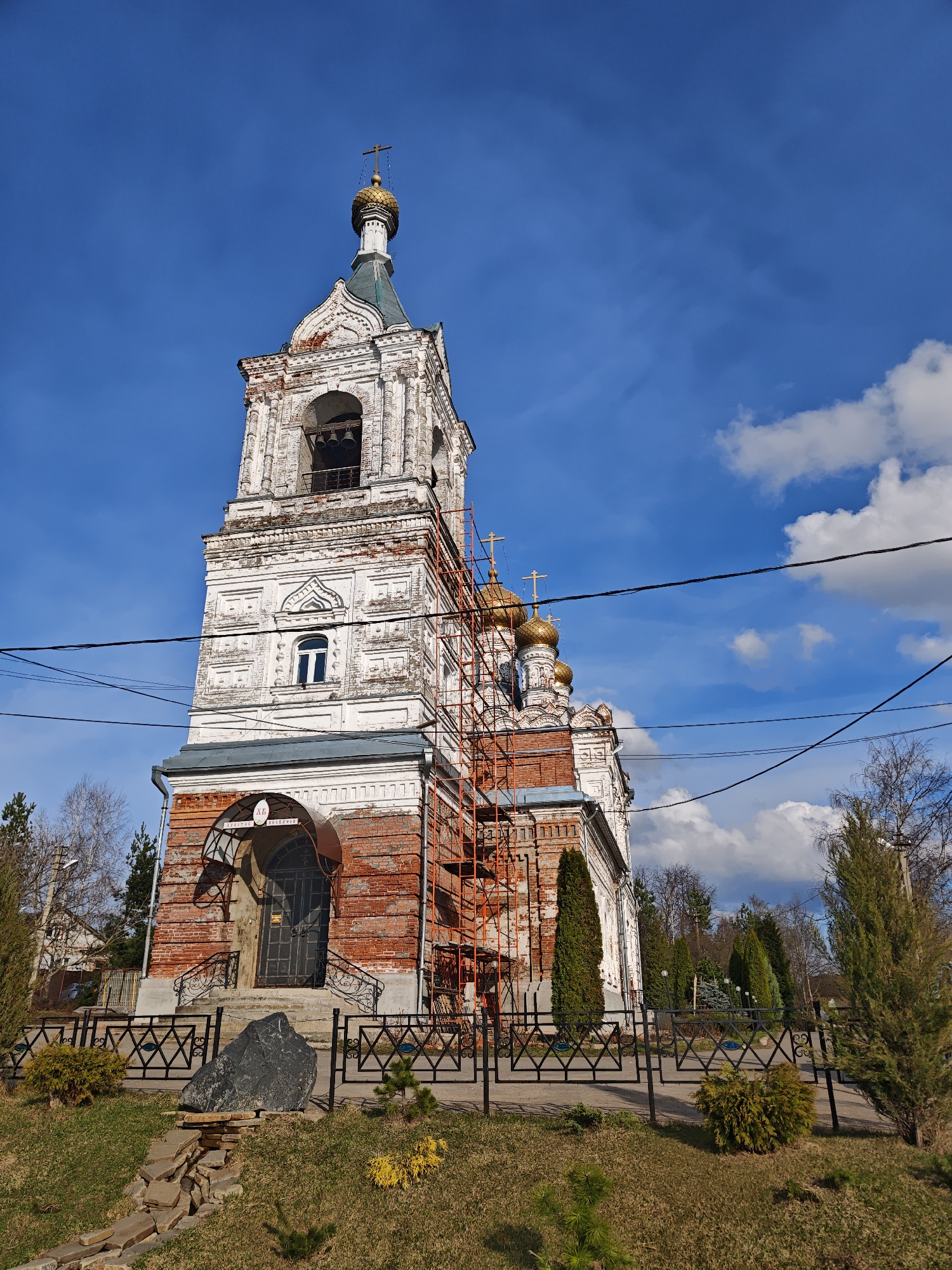 The image depicts the Church of the Life-Giving Trinity in the village of Zhestylevo, Russia. The church, built in the late 18th century, is a tall structure with a prominent bell tower topped with a golden dome and a cross. The exterior of the church is characterized by a mix of white and red brick, showing signs of age and restoration efforts, including scaffolding. The building is surrounded by a metal fence and is set in a landscape of grass, trees, and shrubbery. Power lines traverse the scene. The sky is blue with scattered clouds. The photograph is taken from a low angle, emphasizing the height of the church.