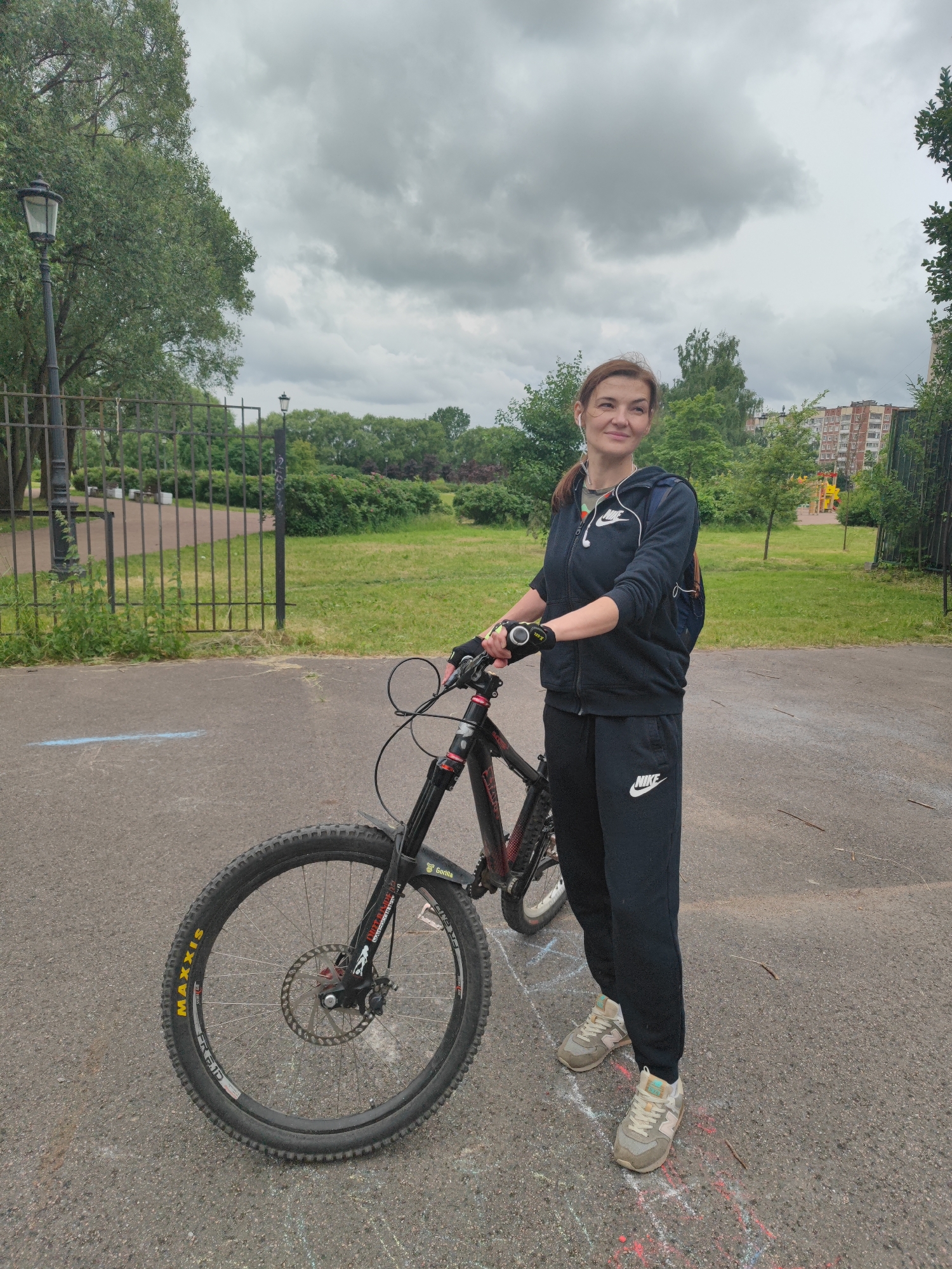 A woman stands next to a bicycle on a paved path in a park on a cloudy day. The woman is wearing a black Nike tracksuit, white earbuds, and light-colored sneakers. She is looking to her right with a slight smile. The bicycle is a dark-colored mountain bike with "Maxxis" written on the tire. In the background, there is a metal fence, trees, green grass, and a multi-story apartment building. The setting appears to be a residential park area in Saint Petersburg, Russia.