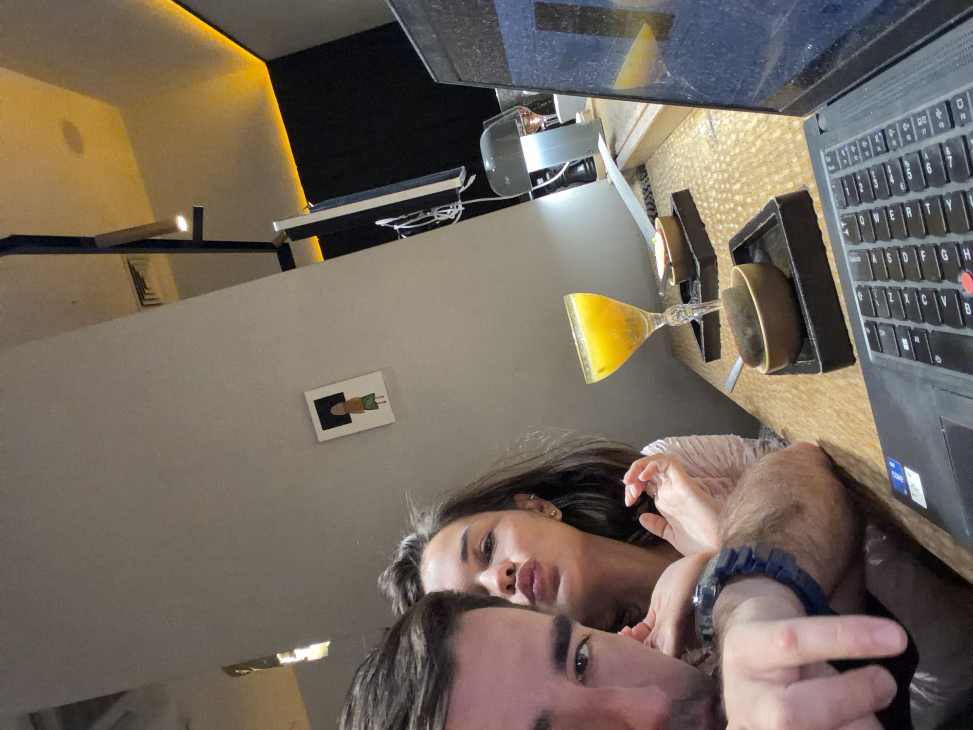 This intimate, rotated image captures a close-up of a couple in what appears to be a cozy indoor setting in Krasnoyarsk, Russia. In the immediate foreground, a man's face is partially visible, primarily his eye and forehead, looking towards the camera. Behind him, a woman's face is clear, looking directly at the lens with a slightly puckered lip expression, suggesting affection or playfulness. Her dark hair falls around her face, and she wears small earrings. The man has a dark watch on his left wrist.
The setting appears to be a modern interior, possibly a bar, restaurant, or a home. To the right of the couple, a black laptop with a QWERTY keyboard is open, displaying stickers for "intel core i7" and "intel iris Xe" on its palm rest. Adjacent to the laptop, on a textured, light-colored surface (resembling woven wood or decorative paneling), are two small dark brown bowls resting in black square holders. A clear glass with a slender stem holds a vibrant orange liquid, likely juice or a cocktail.
The background features warm, indirect lighting emanating from a golden strip along a wall-ceiling joint, casting a soft, ambient glow. A minimalist, rectangular light fixture extends from a wall, and a ceiling vent is visible. On a lighter-colored wall, a small framed picture depicts a stylized figure with a brown top and green bottom against a black background. Various cables and a white power strip are seen on a shelf in the upper left. The overall atmosphere suggests a relaxed, shared moment, likely in the evening.