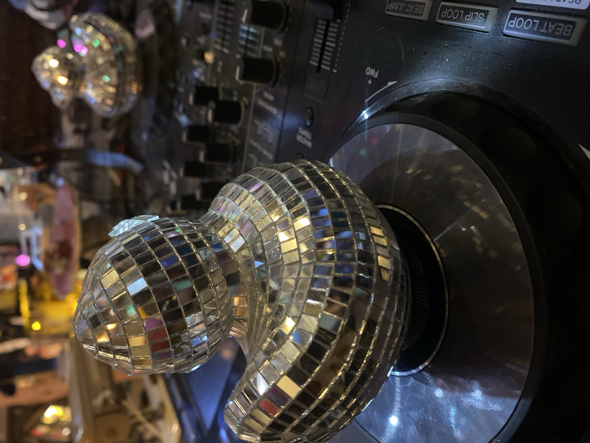 A close-up shot displays a silver, disco-ball-shaped object, attached to a black DJ turntable. The turntable has various buttons and sliders visible, including "BEAT JUMP", "SLIP LOOP", and "BEAT LOOP" labels. A second, blurred disco ball is visible in the background, suggesting an indoor setting, possibly a bar or entertainment venue in Baan Nai Suan, Thailand. The lighting is dim with reflective surfaces, enhancing the disco theme.