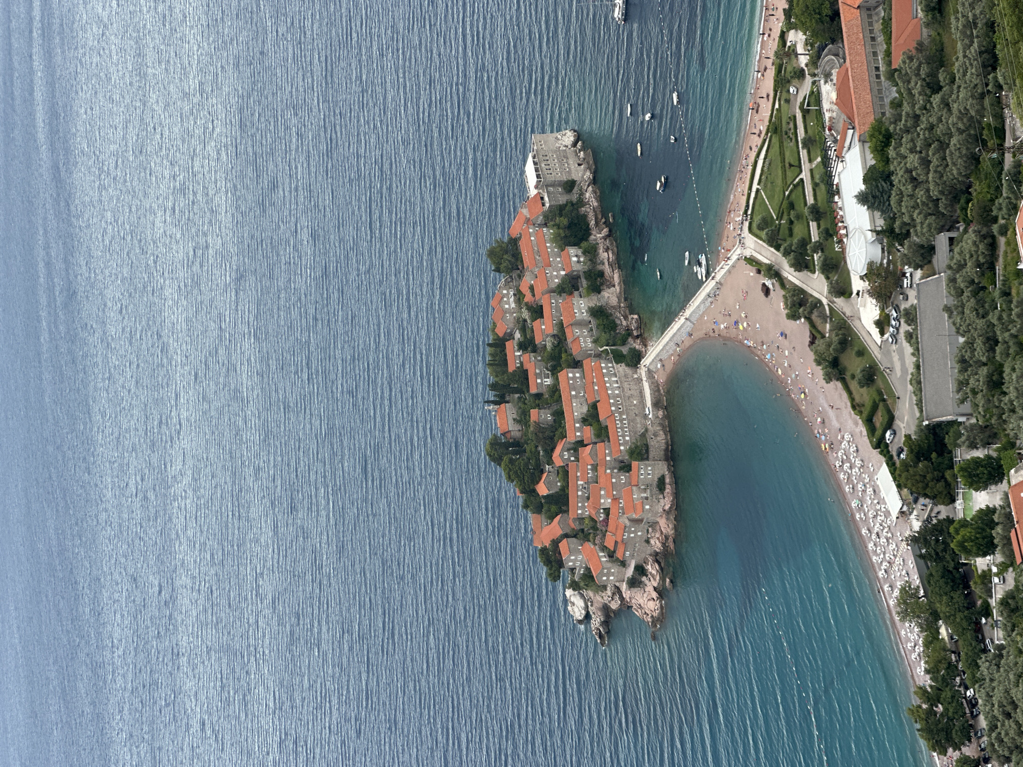 This aerial image captures the picturesque island of Sveti Stefan, Montenegro, during what appears to be a bright, sunny day. The island, connected to the mainland by a narrow causeway, is a charming cluster of red-roofed stone buildings nestled amongst lush green trees. These structures, some appearing to be hotels or residences, are densely packed together on the rocky outcrop, creating a unique architectural landscape.
The surrounding water is a vibrant turquoise, calm and inviting, with numerous small boats dotting its surface. The causeway leads to a sandy beach bustling with activity. Dozens of beachgoers can be seen relaxing on lounge chairs, enjoying the sun, and swimming in the clear sea. A white structure, possibly a beach bar or restaurant, is prominently positioned on the sand.
Adjacent to the beach, well-manicured gardens with pathways wind through the landscape, interspersed with more trees and buildings. The area appears to be a resort or a popular tourist destination. The overall scene evokes a sense of leisure and summer vacation. The image is oriented with the island and coastline occupying the right and bottom portions of the frame, with the vast expanse of the blue sea dominating the left side. No visible text is present in the image.