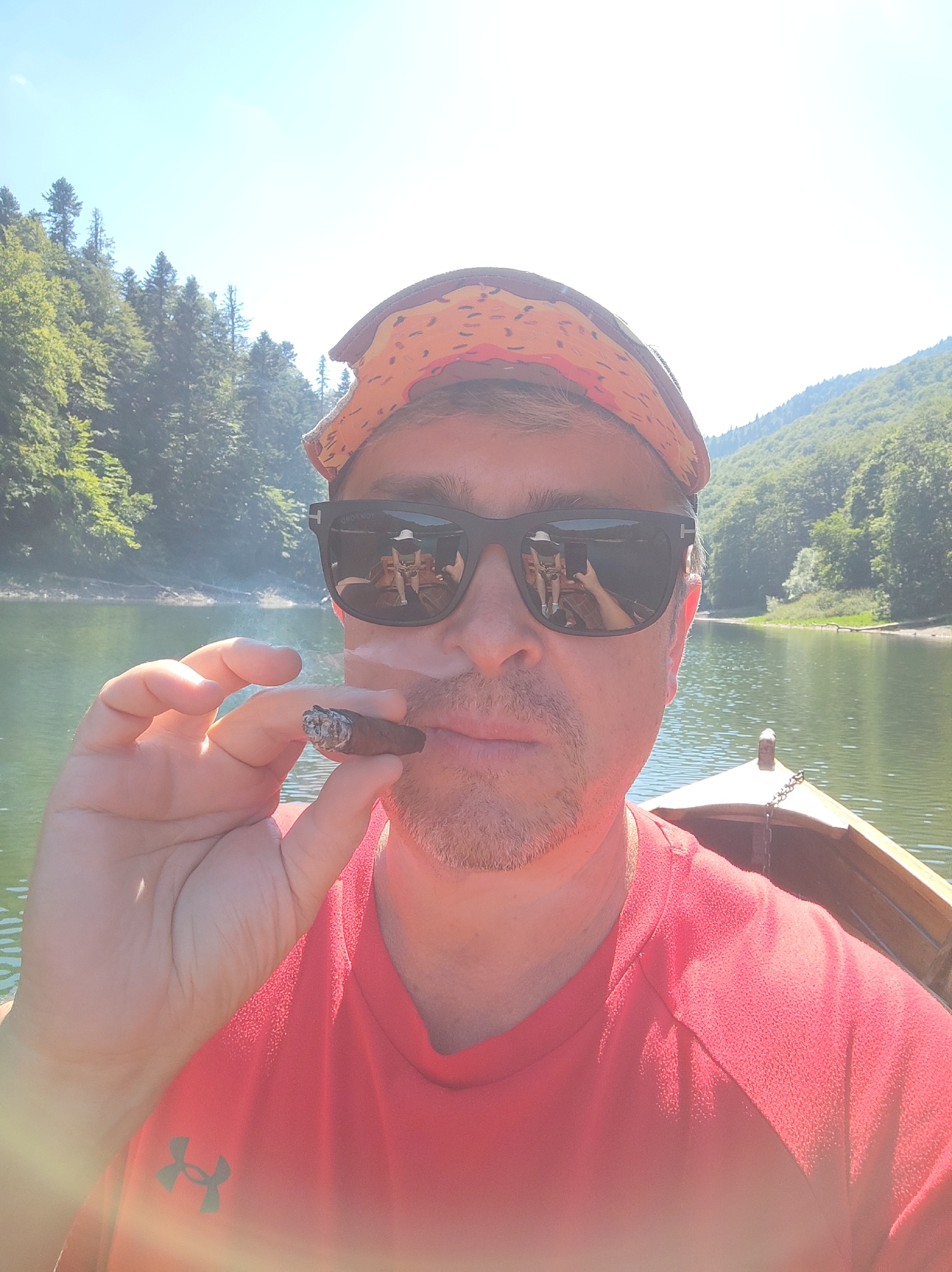In a selfie taken from a boat on a lake, a man wearing sunglasses and a red shirt holds a lit cigar. The lake is surrounded by lush green trees and mountains under a bright blue sky. The man's sunglasses reflect a scene within the boat, showing another person, possibly female, wearing a white hat and a phone. The boat appears to be wooden and is on calm water. The man has stubble and is wearing a patterned bandana. The location is identified as an unknown area in Montenegro.