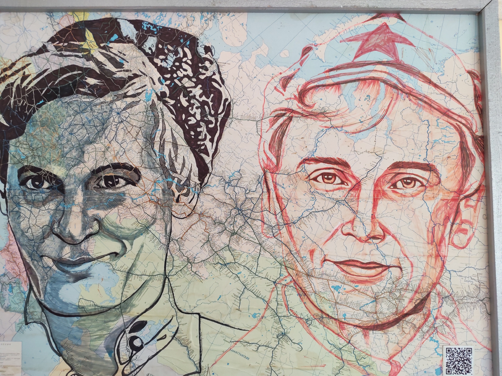 The image shows a framed artwork depicting two portraits overlaid on a map. The left portrait is in shades of gray and black, with details resembling a topological map. The person depicted has short, dark hair and appears to be smiling. The right portrait is rendered in red and light red, with a red star on top of what looks like a hat. The person depicted also appears to be smiling, with lighter skin tones. The underlying map shows various lines, waterways, and place names written in Cyrillic script. A QR code is visible in the bottom right corner. It is stated to be located in the city of Agryz, Russia.
