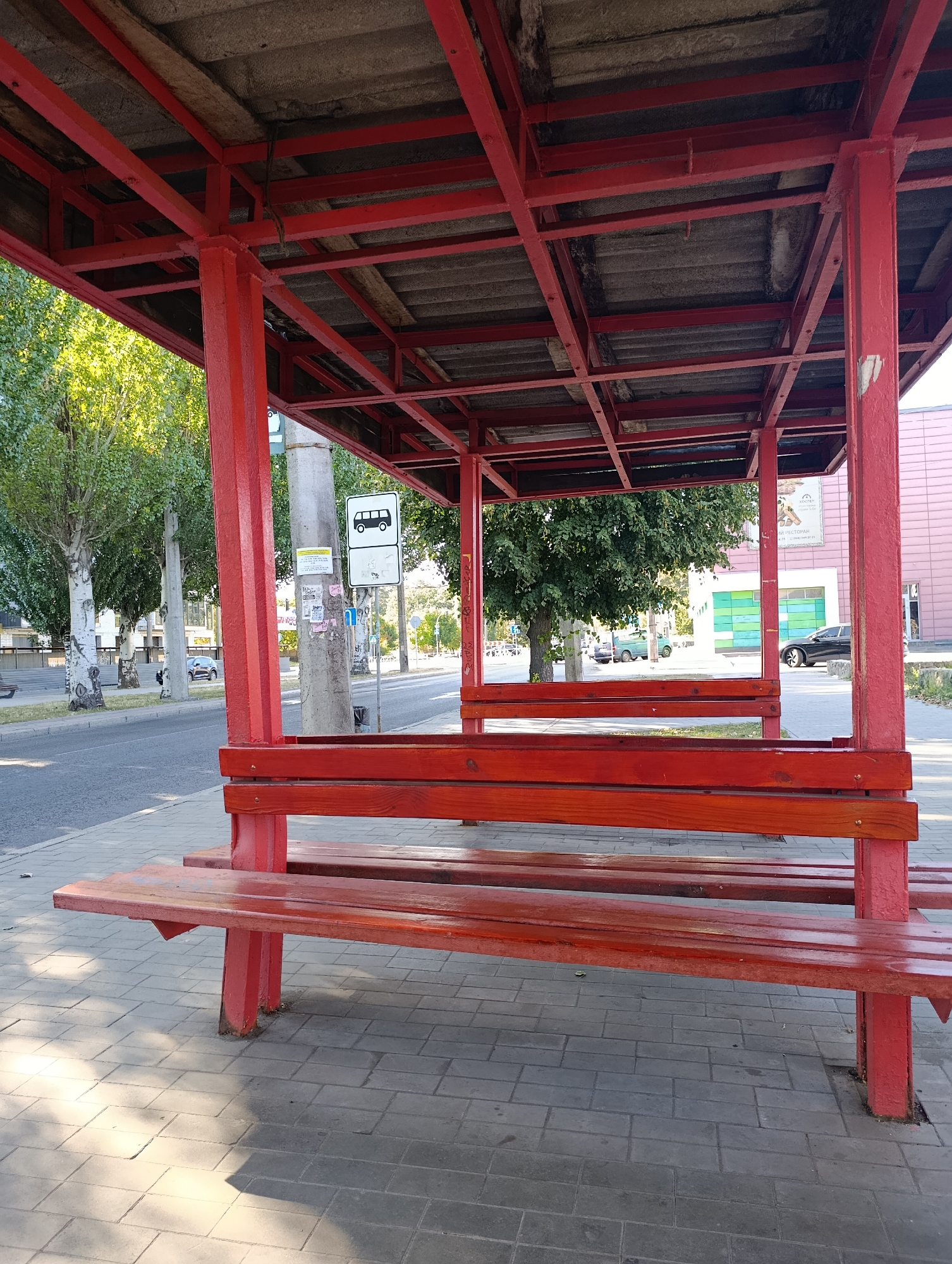 The image depicts a bus stop shelter with a red metal frame and wooden benches, situated on a paved sidewalk next to a street. Trees line the street, and buildings are visible in the background. A bus stop sign is mounted on a pole near the shelter. The scene appears to be a typical urban environment during the daytime, with no people or vehicles in the immediate vicinity of the bus stop.