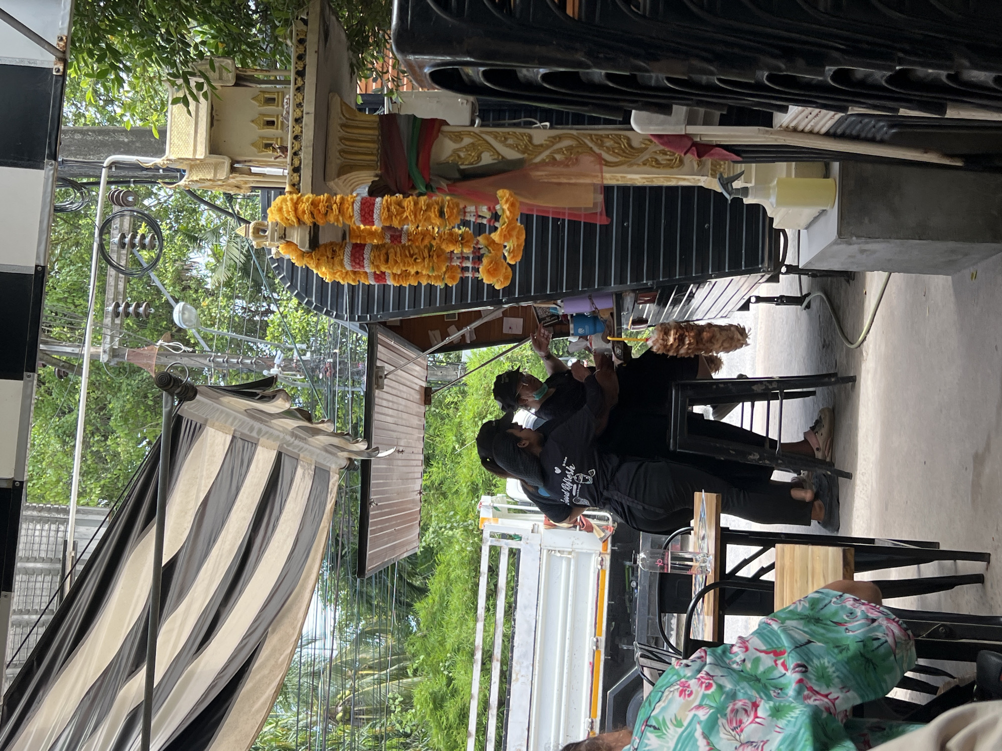 In Choeng Thale, Thailand, a street-side food stall operates under a striped awning, with a white truck visible in the background. Two women in black shirts and pants are working at the stall, one appearing to prepare food or drinks. They are interacting with each other and possibly a customer. A small, ornate shrine adorned with yellow marigold garlands stands to the right of the stall. Stacks of black chairs are visible near the shrine, and a person wearing a green floral shirt is seated at a wooden table in the foreground, looking away from the camera. The scene suggests a casual, outdoor dining or service environment, possibly on a sunny day given the lighting.