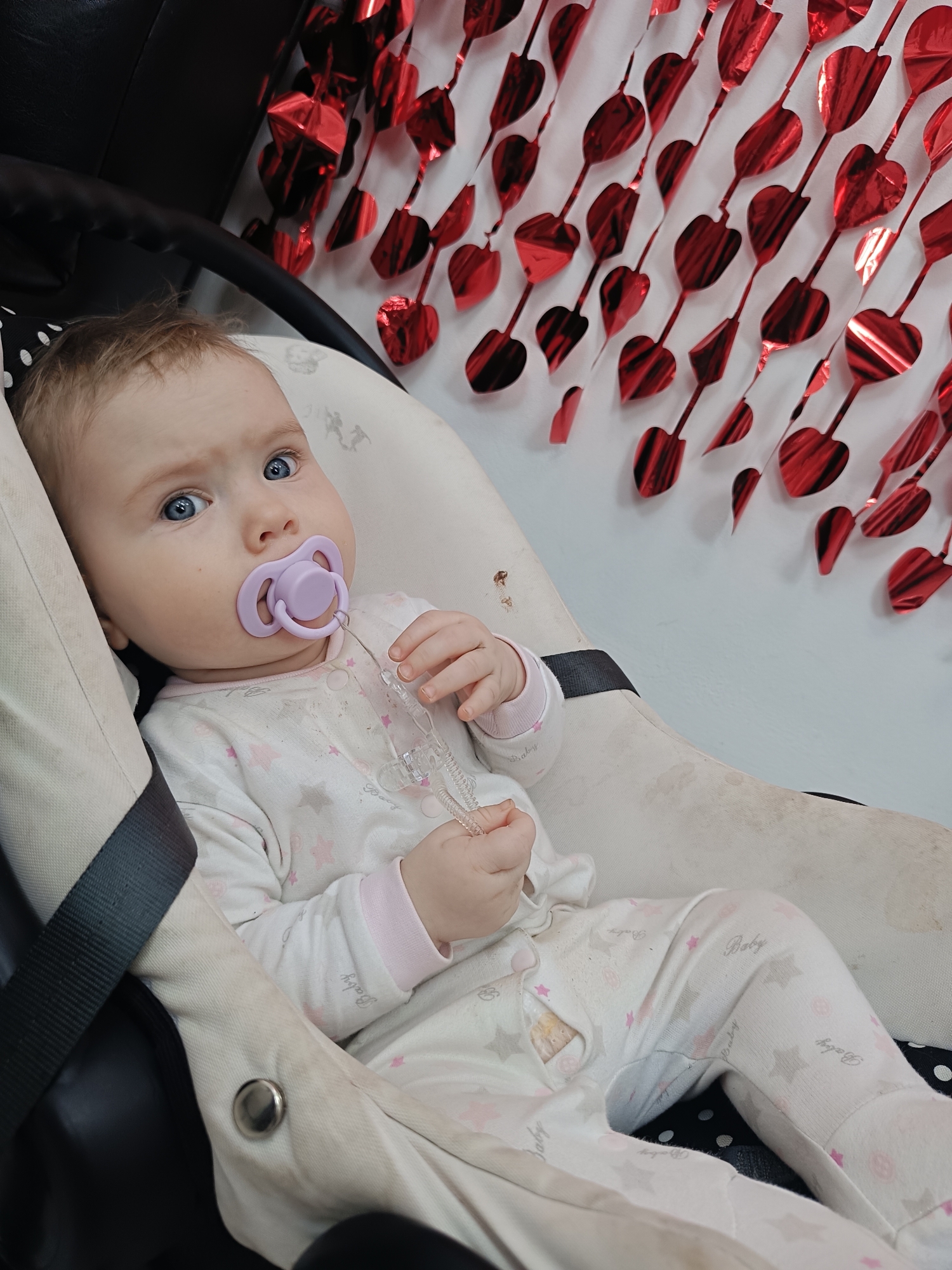 The image features an infant with fair skin, light hair, and blue eyes, sitting in a beige baby seat or car seat insert. The baby looks directly at the camera with a calm, observant expression. A purple pacifier is in the baby's mouth, and a clear plastic pacifier clip is held in one hand.
The baby is dressed in a white sleepsuit or onesie patterned with pink and grey stars, and the word "Baby" is printed repeatedly on the fabric. The beige fabric of the baby seat and parts of the baby's clothing show some minor discoloration or stains. Black straps of the seat are visible, securing the baby.
The background consists of a plain white wall adorned with a vertical decoration of shiny red metallic heart shapes, strung together like a garland. This festive element fills the upper right portion of the frame. The scene appears to be indoors, likely during the day due to ample lighting. No specific outdoor conditions are discernible.