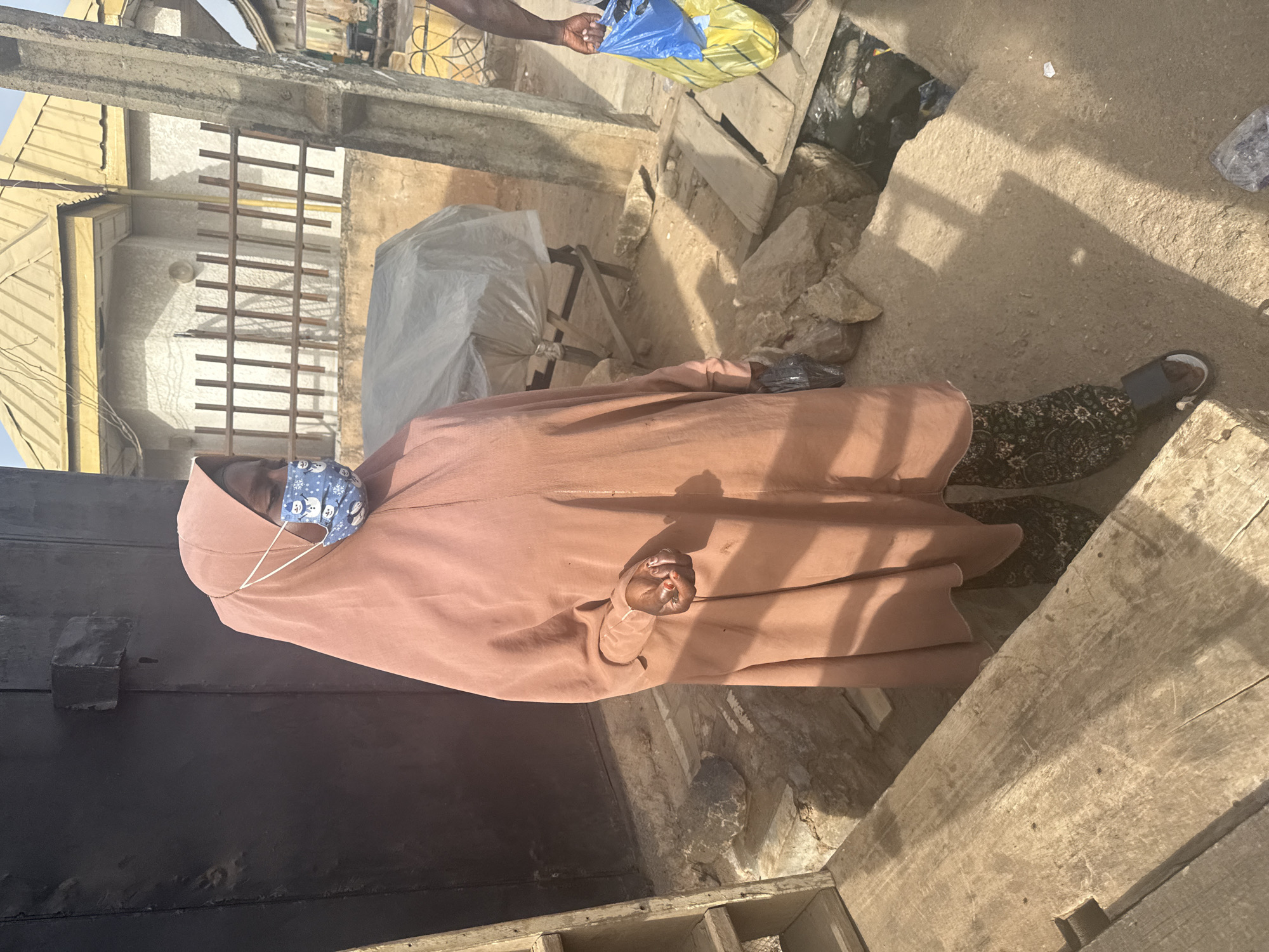 A woman wearing a light brown hijab and a patterned face mask stands in an urban environment in Jos, Nigeria. She also is wearing pants with a dark paisley pattern. Her left foot is visible with a brown sandal on it. The area around her appears to be an alley or narrow space between buildings with a concrete or dirt ground. There is a person in the upper right of the image, visible from the waist up, holding a plastic bag in blue, yellow and transparent colors. There is trash on the ground. To the left of the woman is a dark door or wall made of wooden planks. There is a building with a metallic roof and a partially covered object wrapped in white plastic.