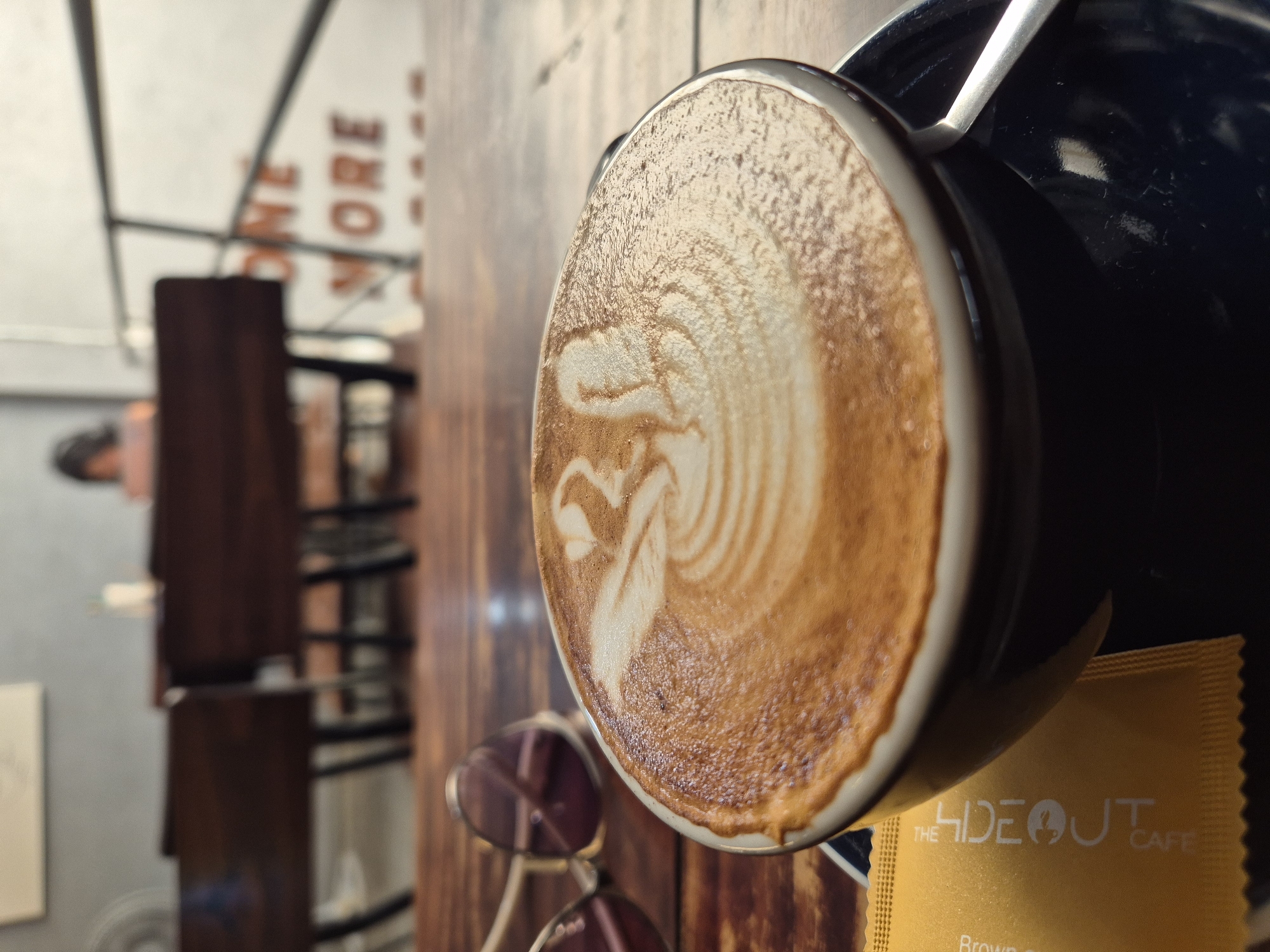 The image shows a close-up of a cup of latte art with a swan design on top, placed on a dark saucer on a wooden table. To the left of the cup, a pair of sunglasses with purple lenses and gold frames is visible. A yellow sugar packet with the text "THE HIDEOUT CAFE" is placed under the cup. In the background, blurred wooden chairs and tables are arranged, suggesting a cafe setting. A person is partially visible in the far background, seated at a table. The overall lighting suggests an indoor environment with natural light. The text "ONE MORE" is visible on a wall in the background, which is a common phrase found in cafes. The cafe "The Hideout Cafe" is a known establishment in Da Nang, Vietnam, often frequented by locals and tourists for its coffee and ambiance.